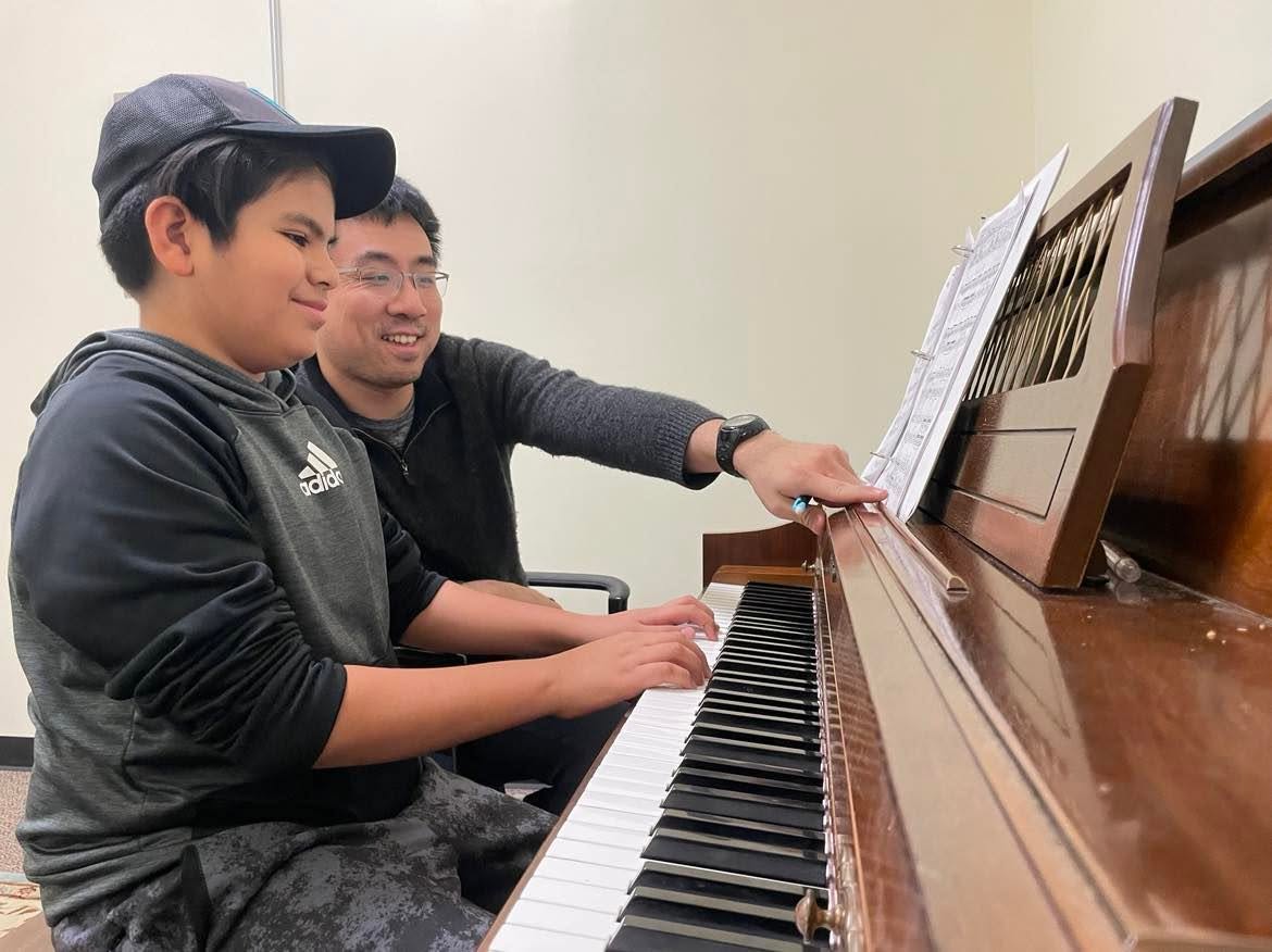 Two people, a young boy and an adult, sit at a piano, smiling and looking at sheet music, with the boy playing the keys.