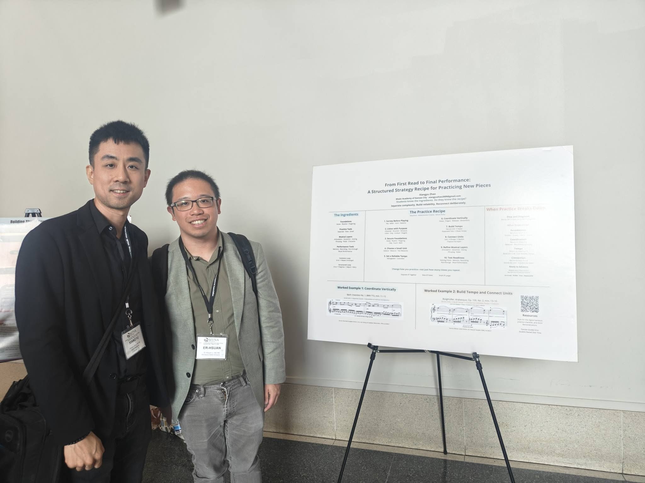 Two people standing next to a large presentation poster on a stand, smiling, in a plain room.