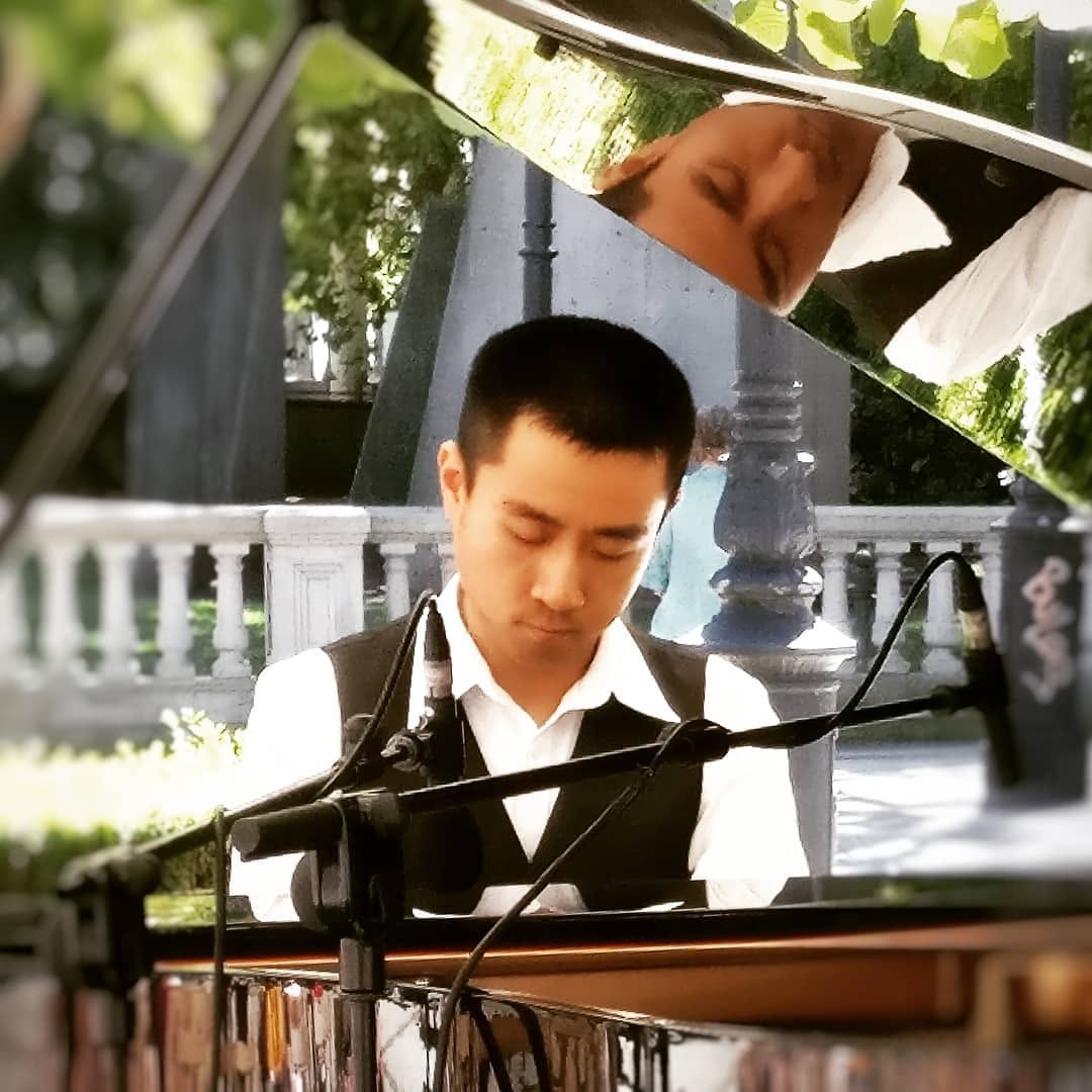 Young boy playing piano outdoors, focused on keys, with trees and a fence in background.