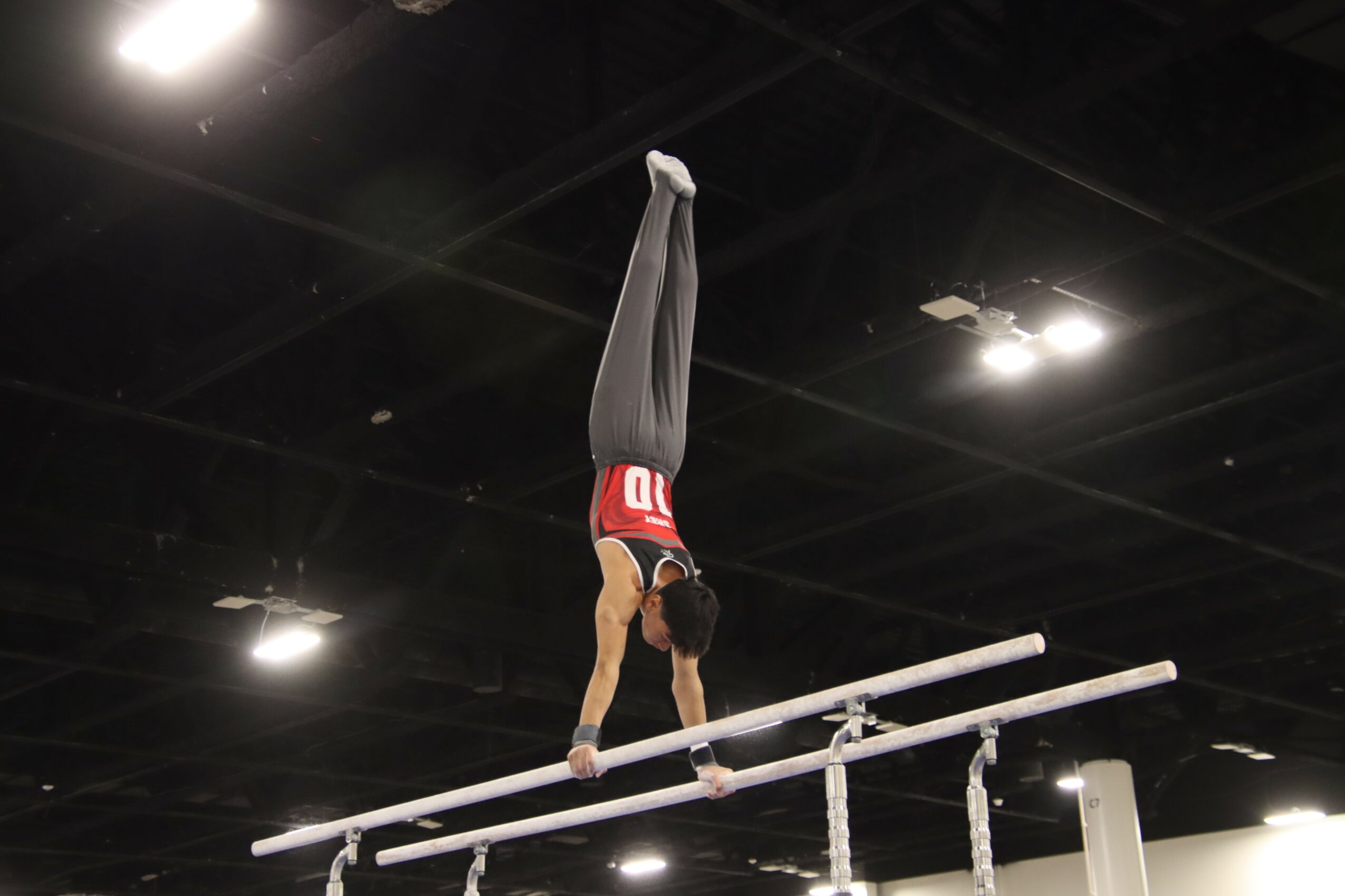 Person performing a handstand on parallel bars in an indoor gym, with ceiling lights visible above.