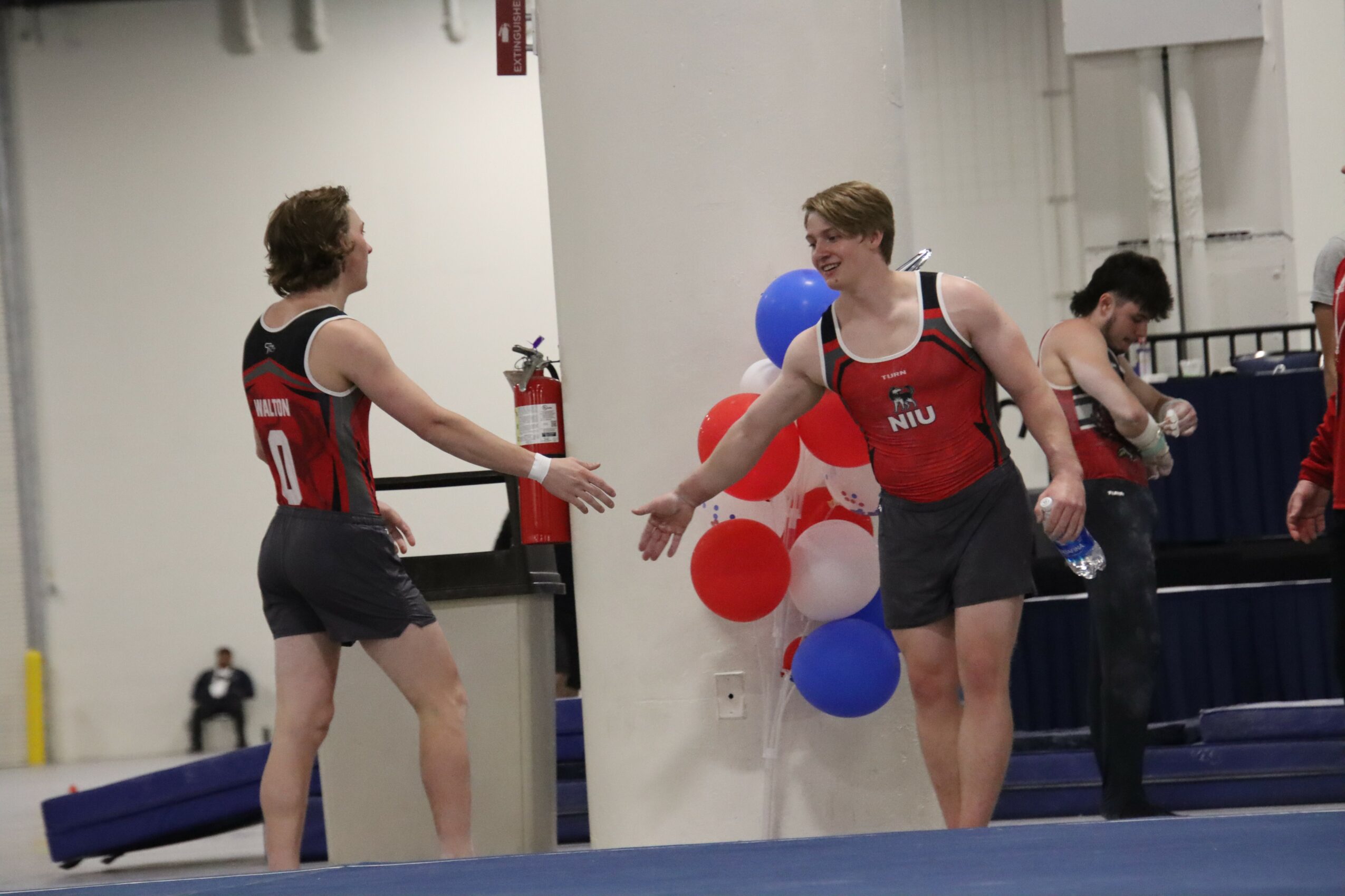 Two athletes in red and black uniforms shake hands in a gymnasium, with balloons and a person sitting nearby.