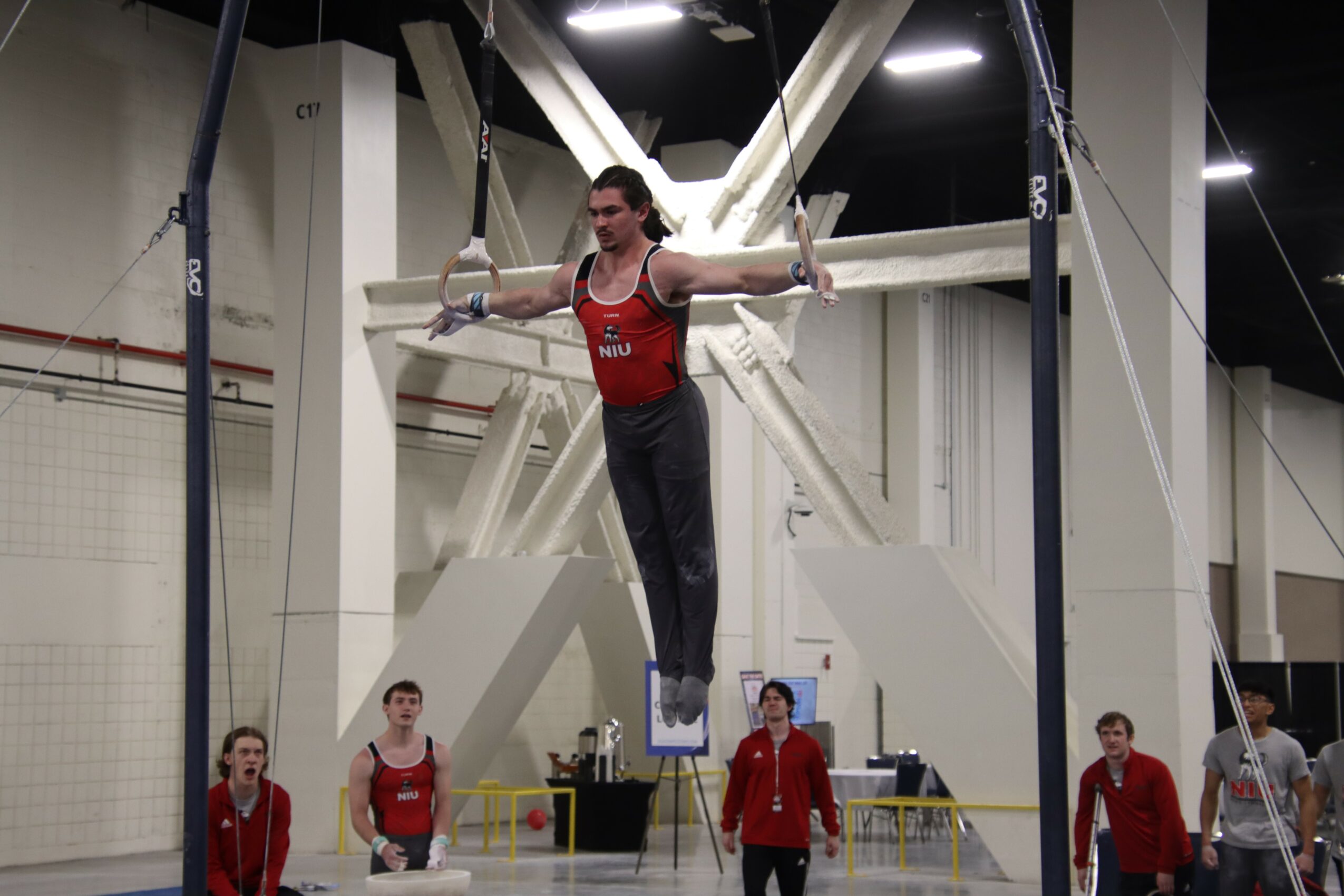 Female gymnast on uneven bars in an indoor gymnasium, wearing a red and black leotard, with other gymnasts nearby.