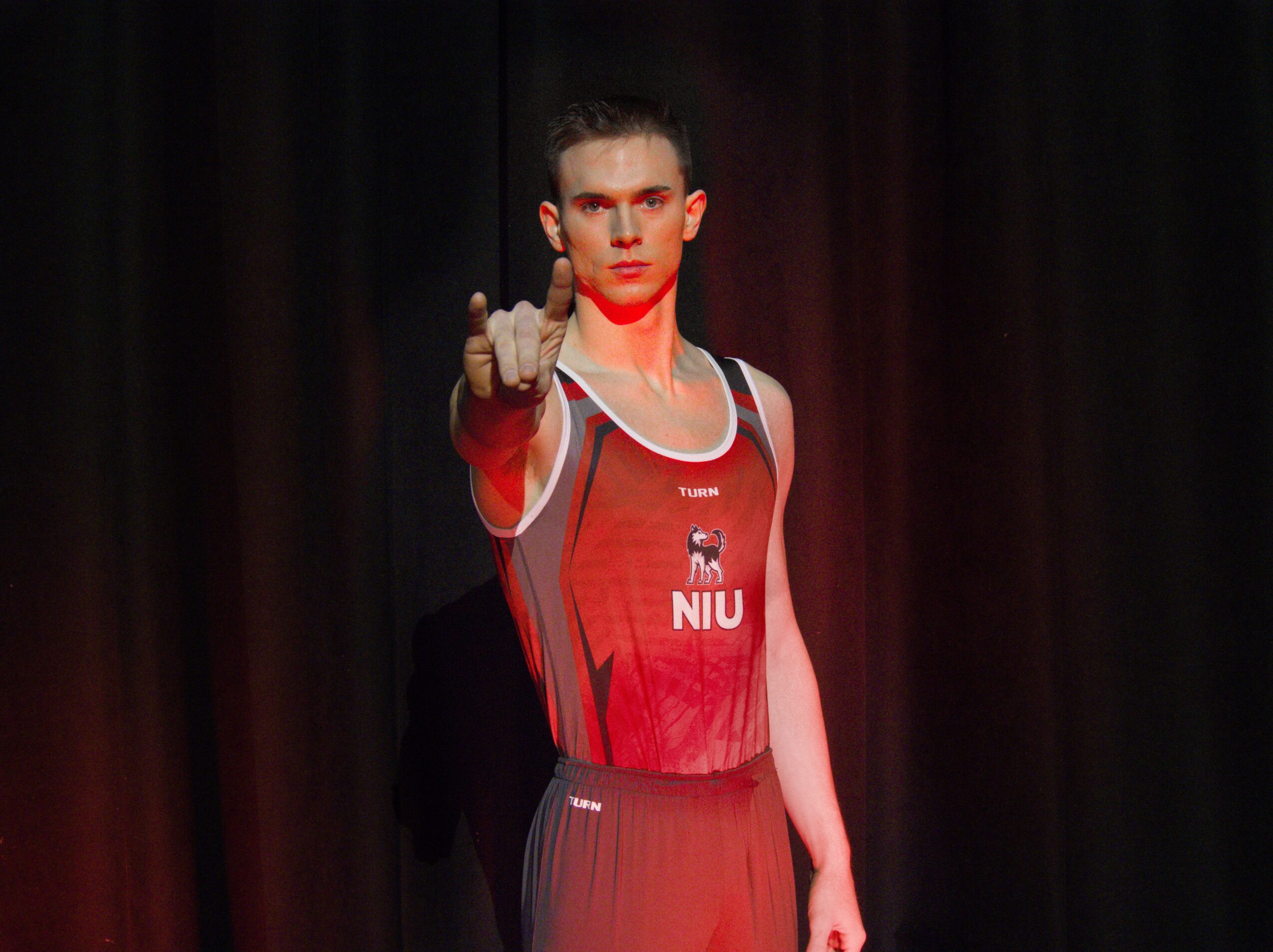 Person in athletic attire pointing forward, standing on stage with dark background.