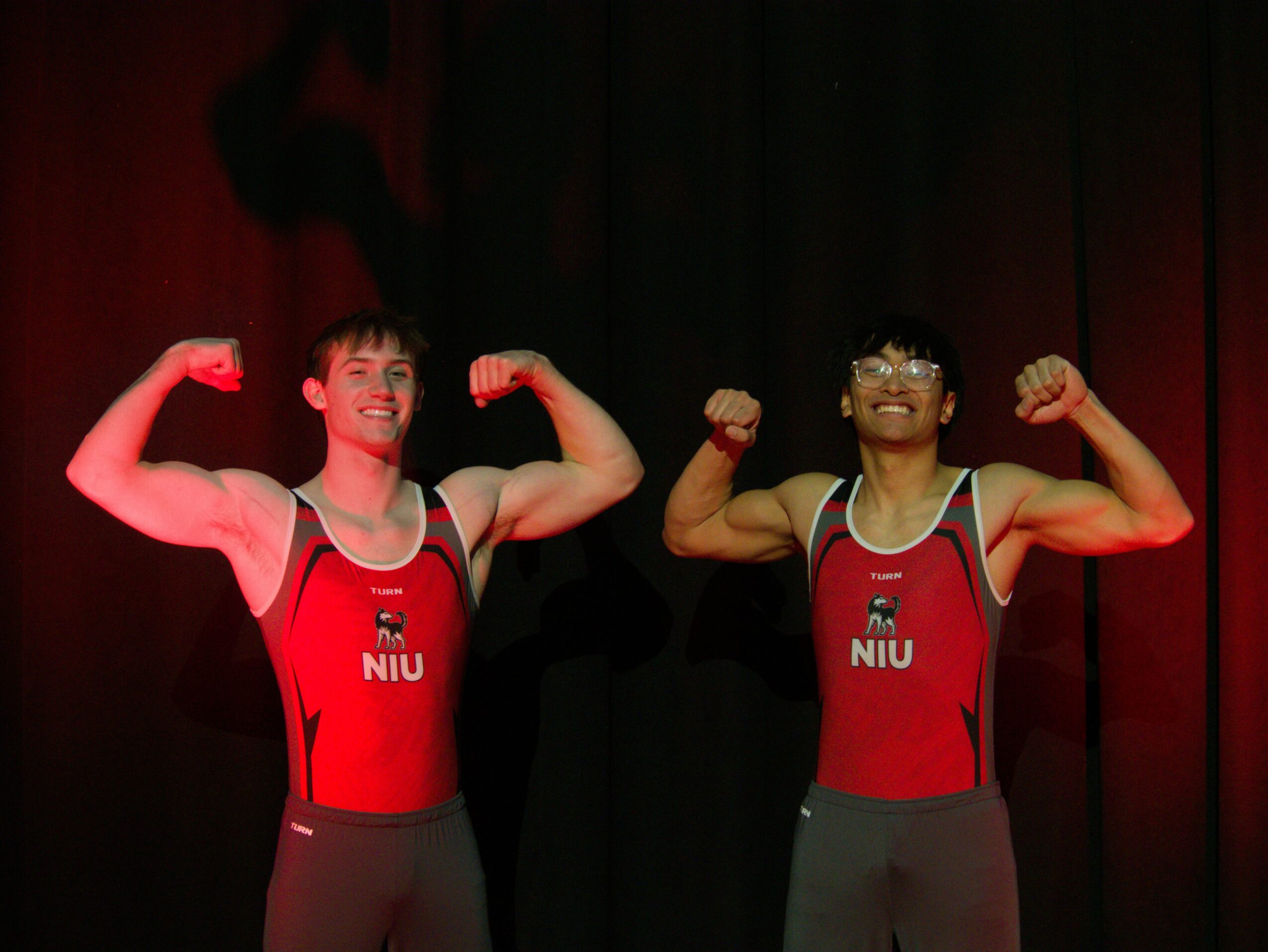Two athletes in red uniforms flexing their arm muscles on stage with a dark background.