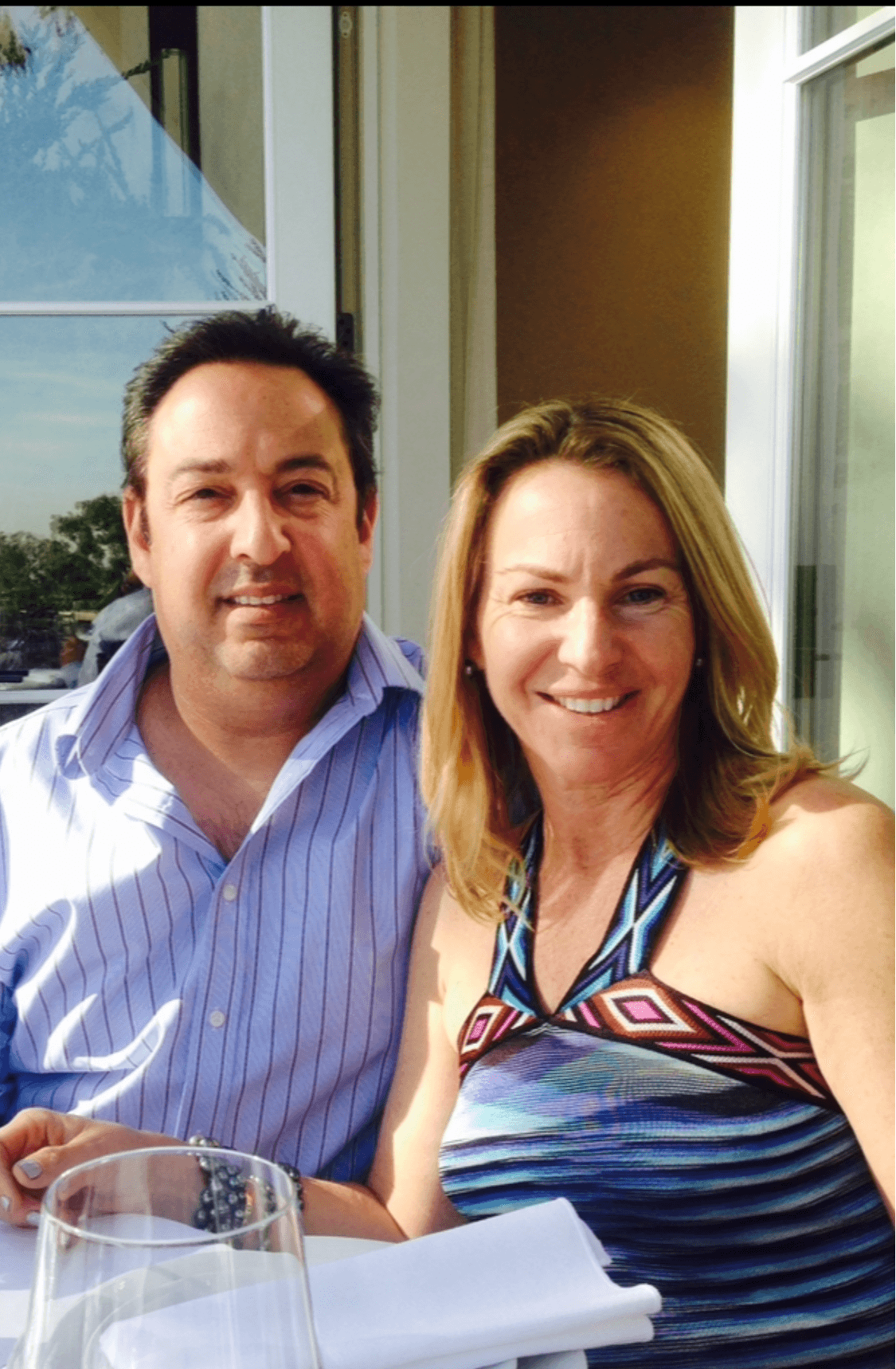 Two people smiling indoors near large windows, with a glass and napkin on a table in front.