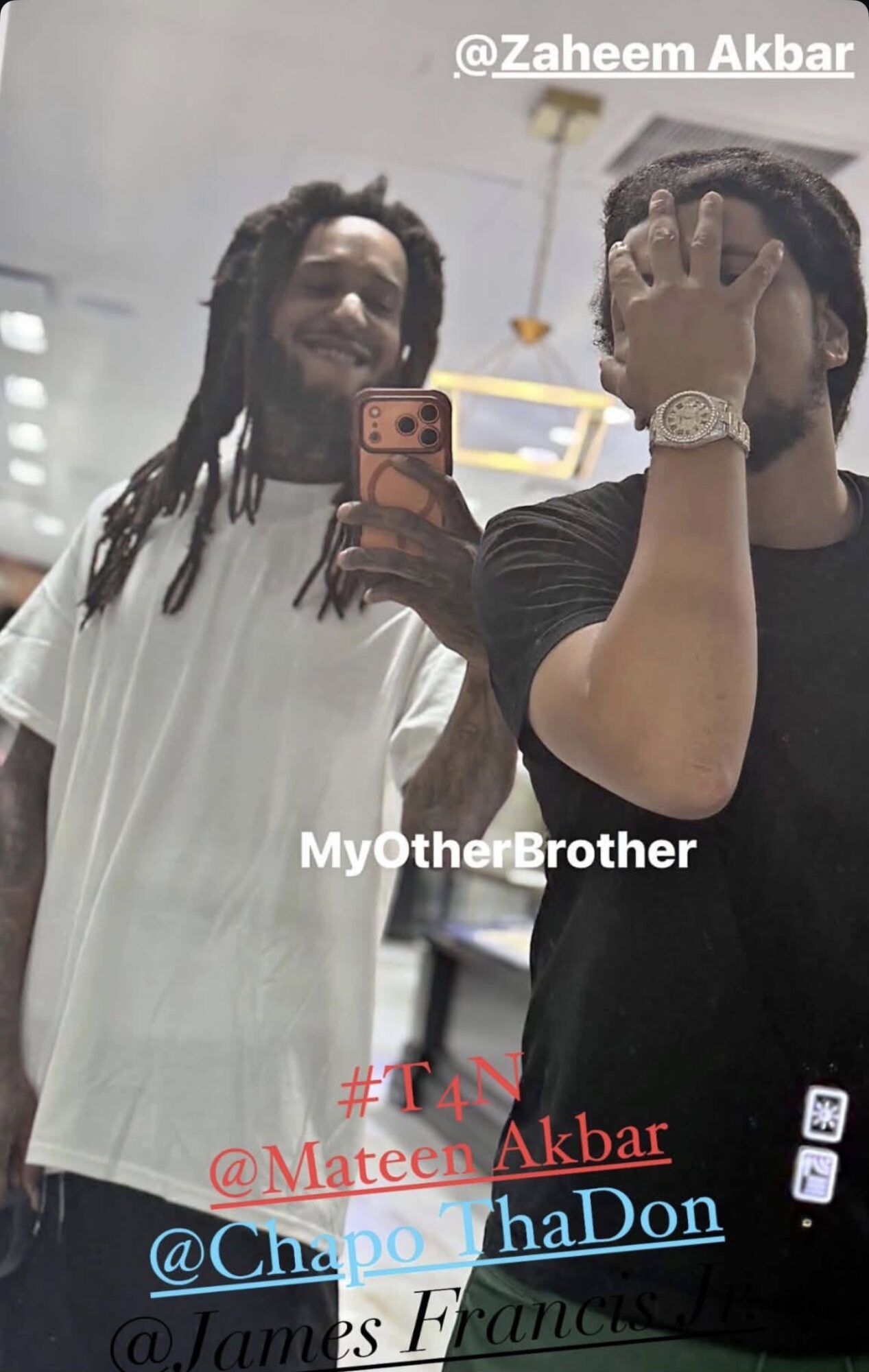 Two men taking a mirror selfie, one with dreadlocks smiling, the other covering face with hand, in a bright room.