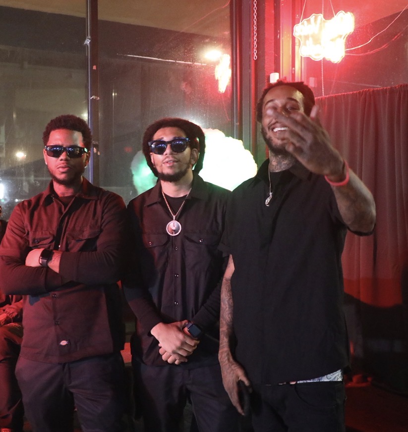 Three men standing indoors, two wearing sunglasses, one smiling and covering his mouth, with red and green lighting in the background.