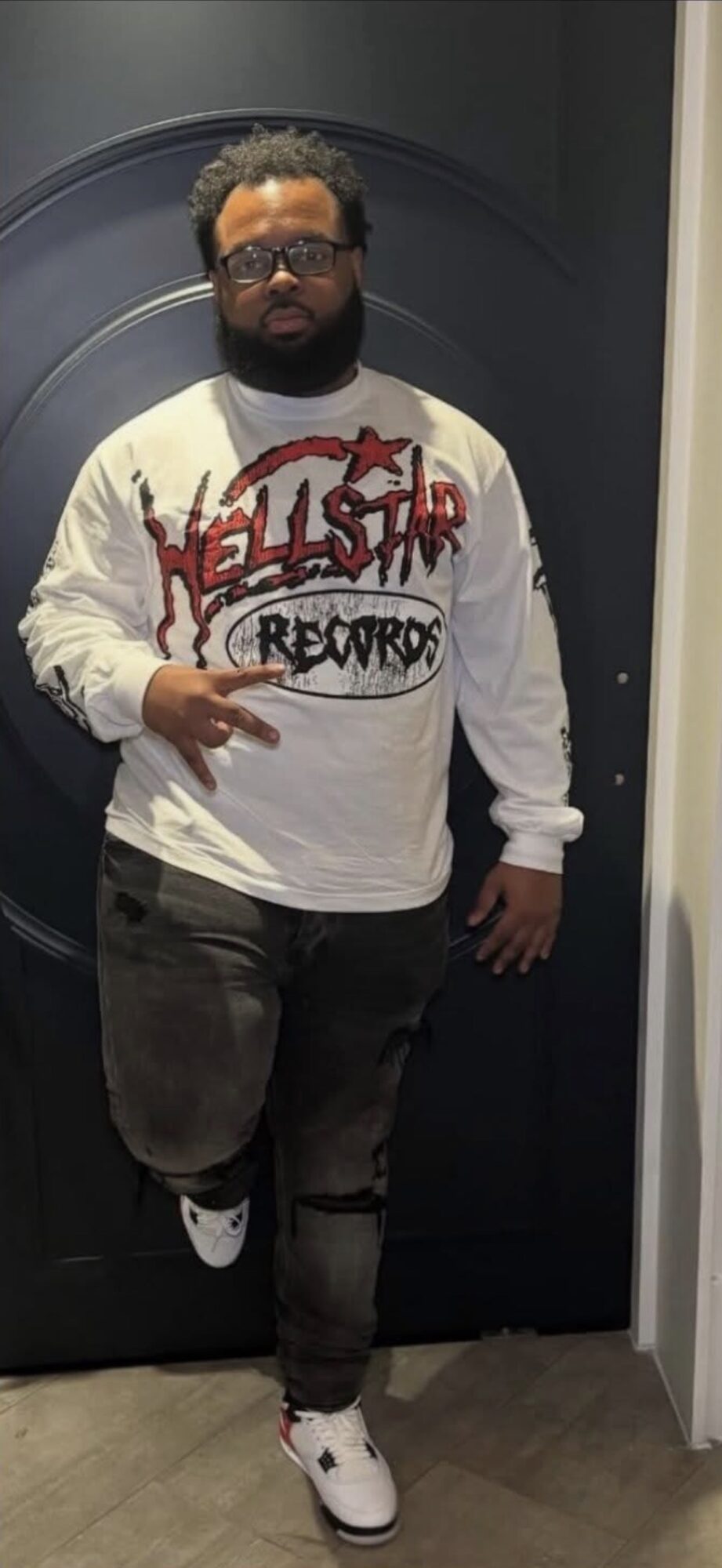 Man with glasses and beard standing in front of a dark door, wearing a white sweatshirt with red and black text, black pants, and white shoes.