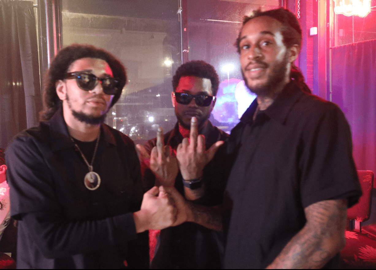 Three men standing together in a dimly lit setting, two wearing sunglasses and making hand gestures, one smiling at the camera.
