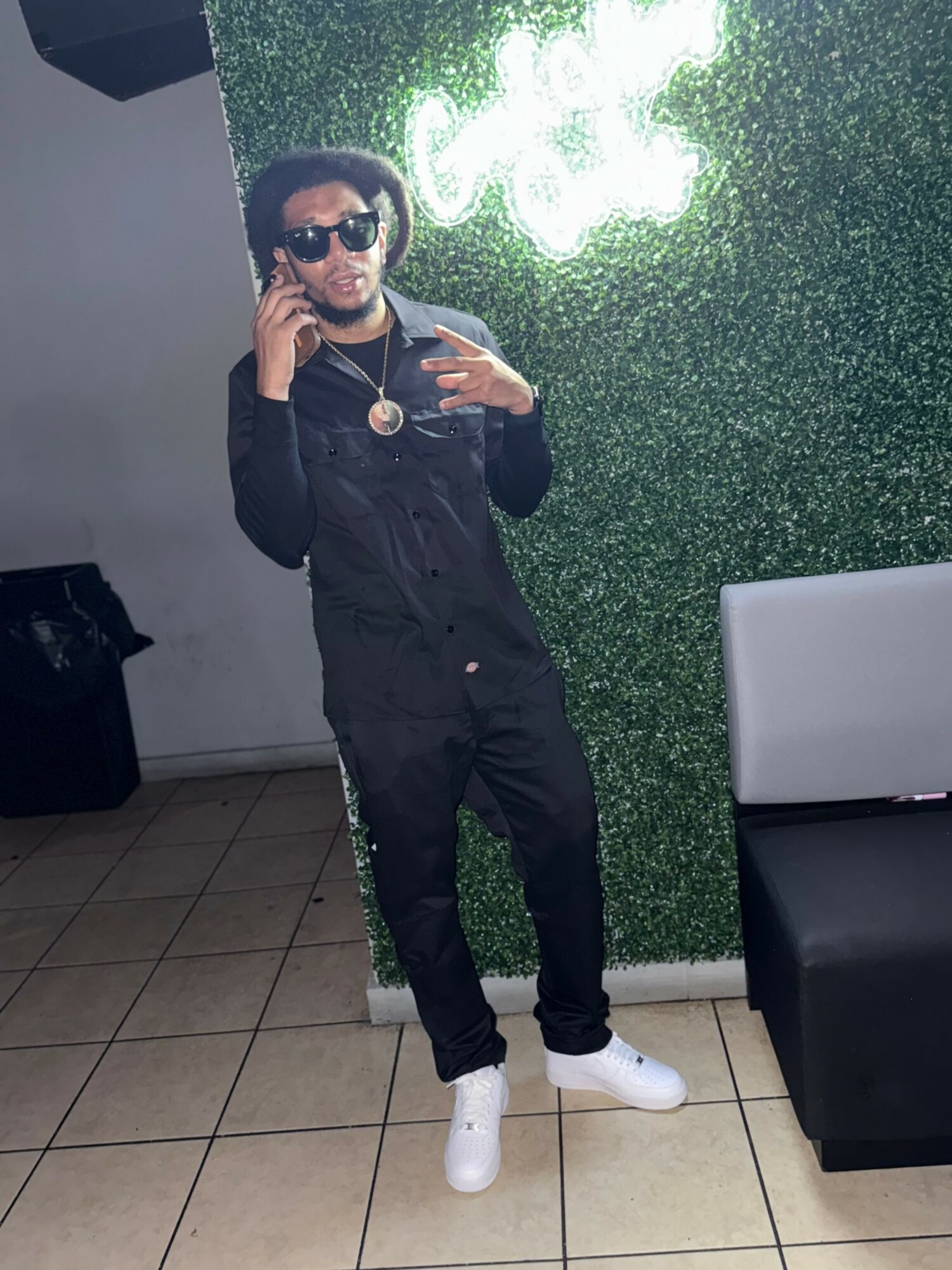 Person taking a mirror selfie, wearing sunglasses, black outfit, white shoes, and a hat, standing in front of a green wall with a neon sign.