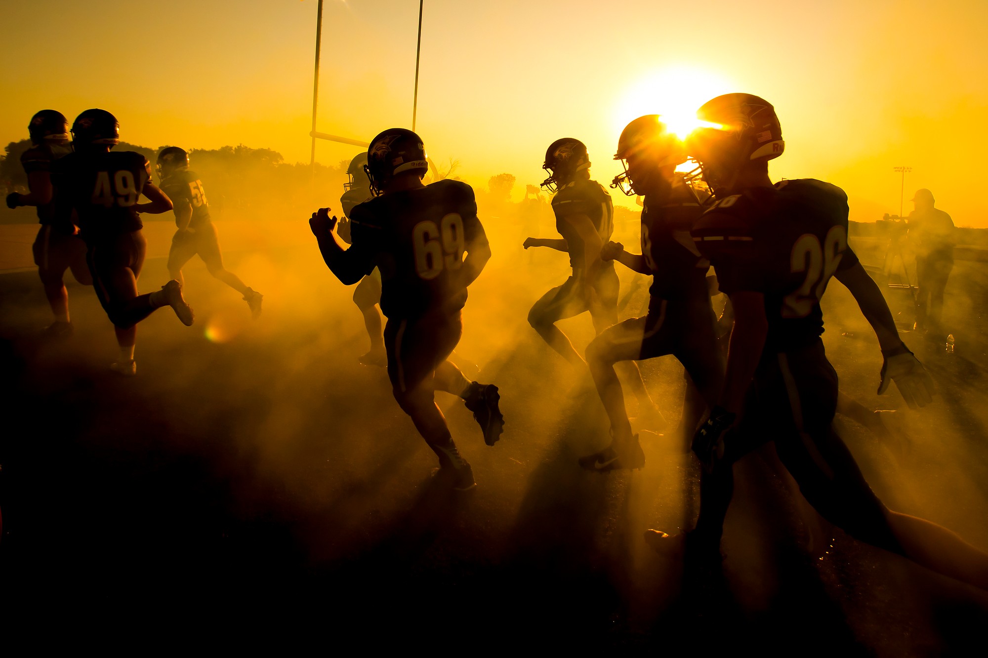 Football players running on a field with dust and a bright sunset in the background.