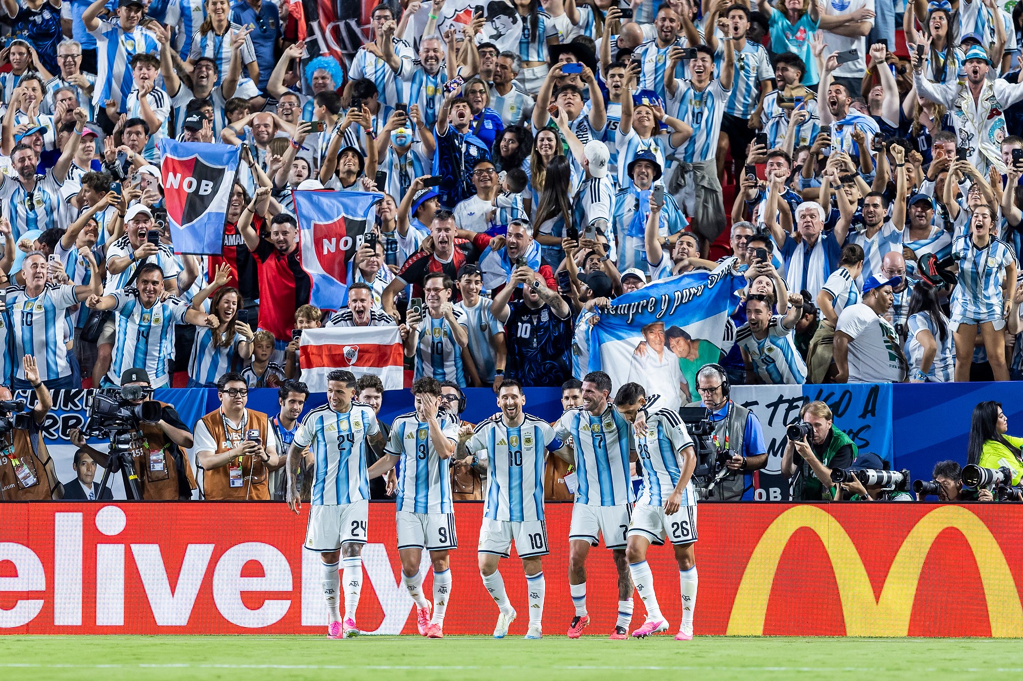 Group of soccer players in blue and white striped uniforms standing on field, with crowd in background holding flags and banners.