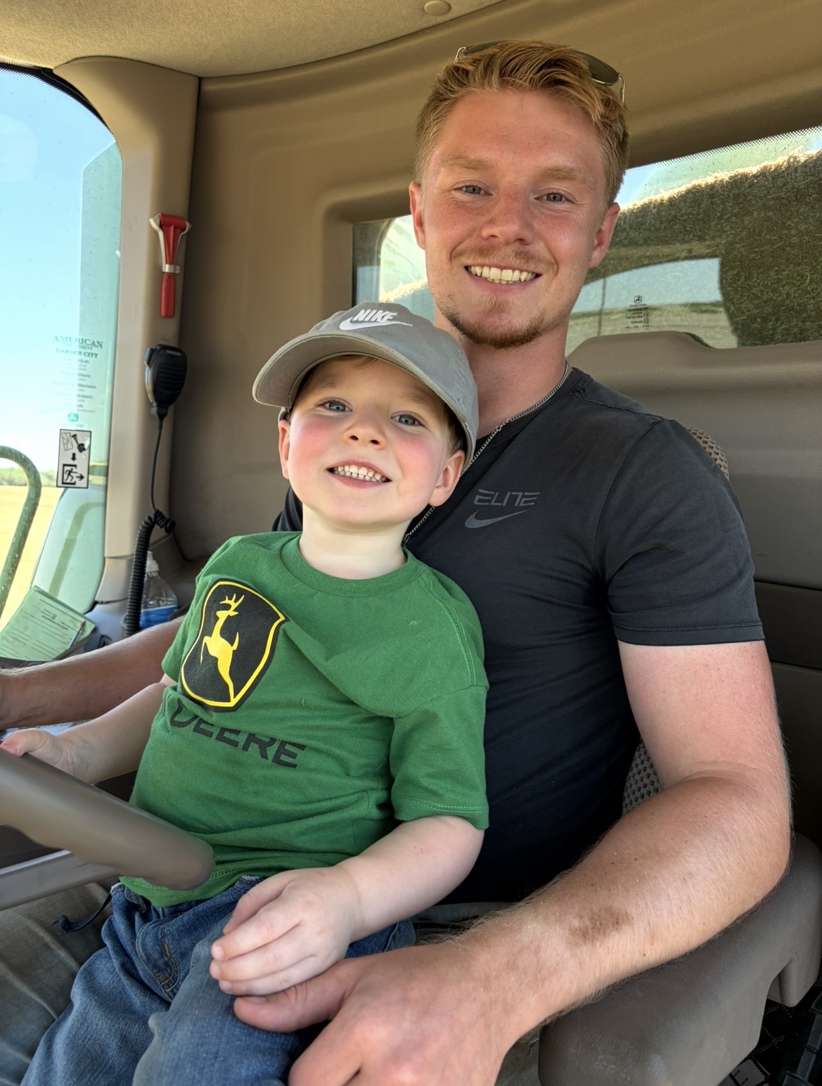 A young man and a child sitting inside a vehicle, smiling at the camera. The child wears a green shirt and gray cap.