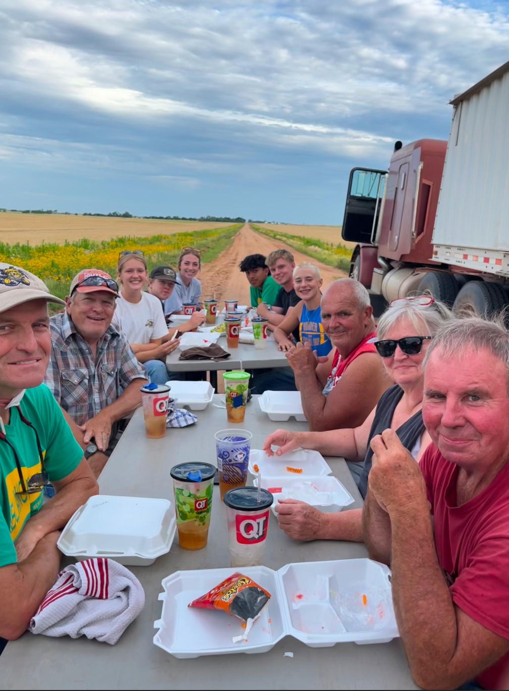 Group of people sitting at a long outdoor table with food and drinks, countryside background, truck nearby, sunny sky.