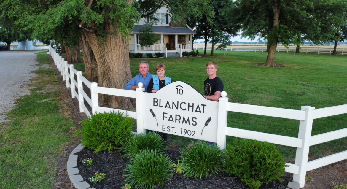 Three people standing behind a white fence near a large tree and a sign for Blanchard Farms, established 1902.