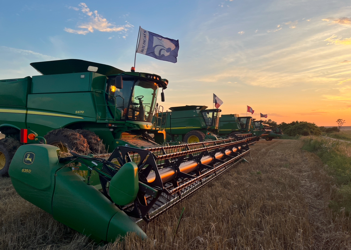 Three green harvesters with flags on a field during sunset, facing right, with a clear sky and horizon.