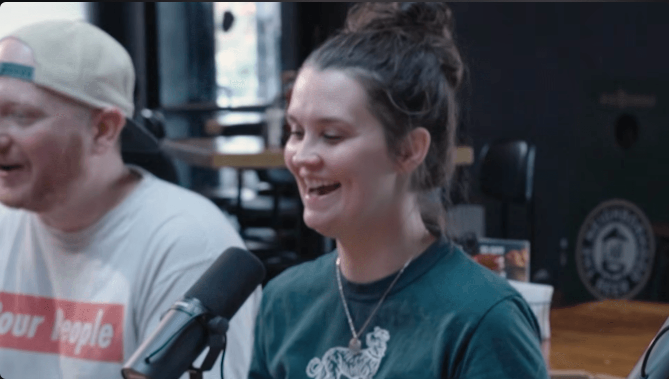 Two people smiling and speaking into a microphone in a room with dark walls and wooden furniture.