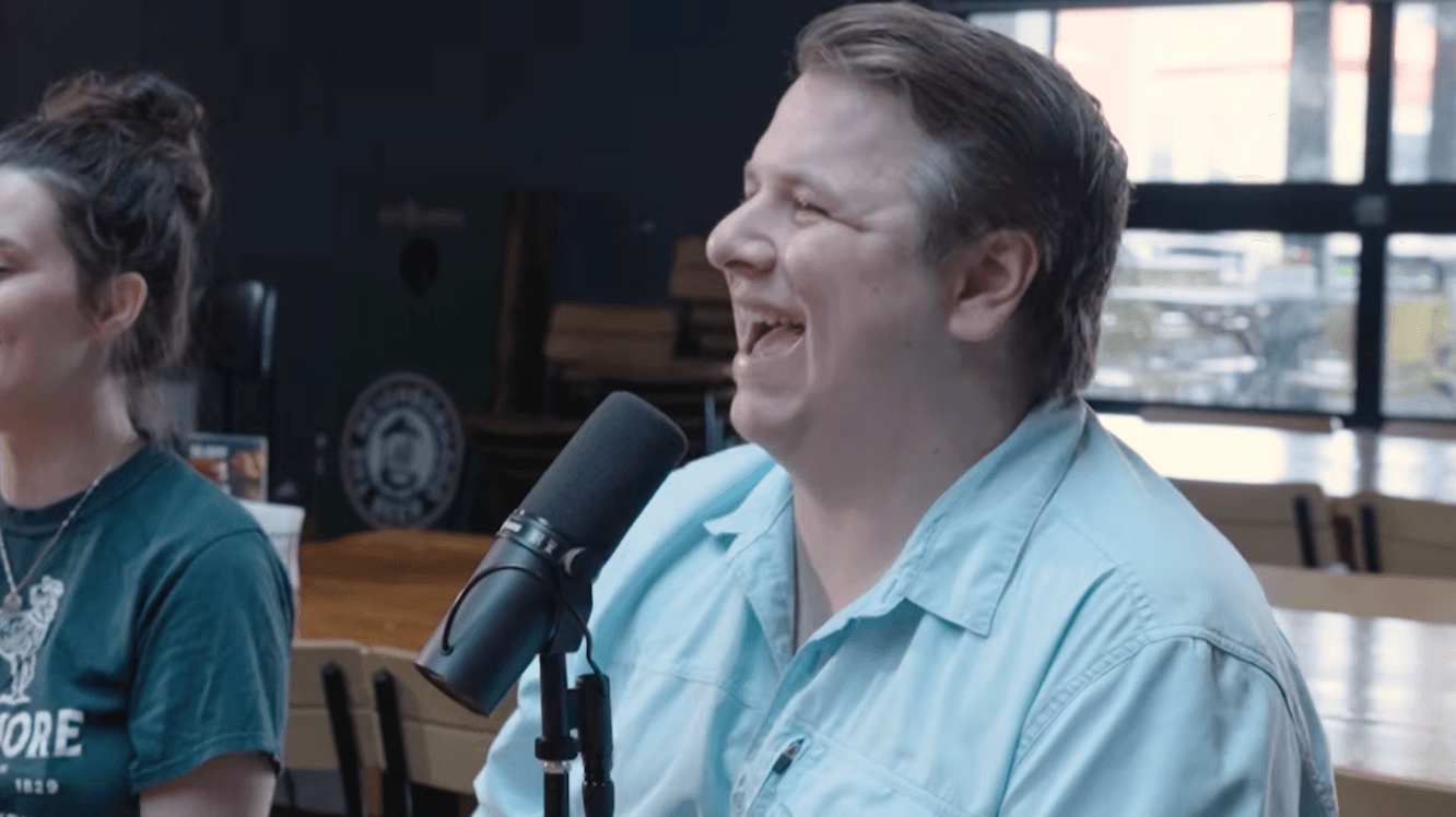 Man laughing into microphone during recording session, woman partially visible beside him, indoor setting with large windows