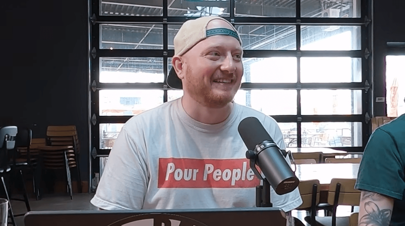 Smiling man with a beard and cap, sitting at a table with a microphone, in a room with large windows.