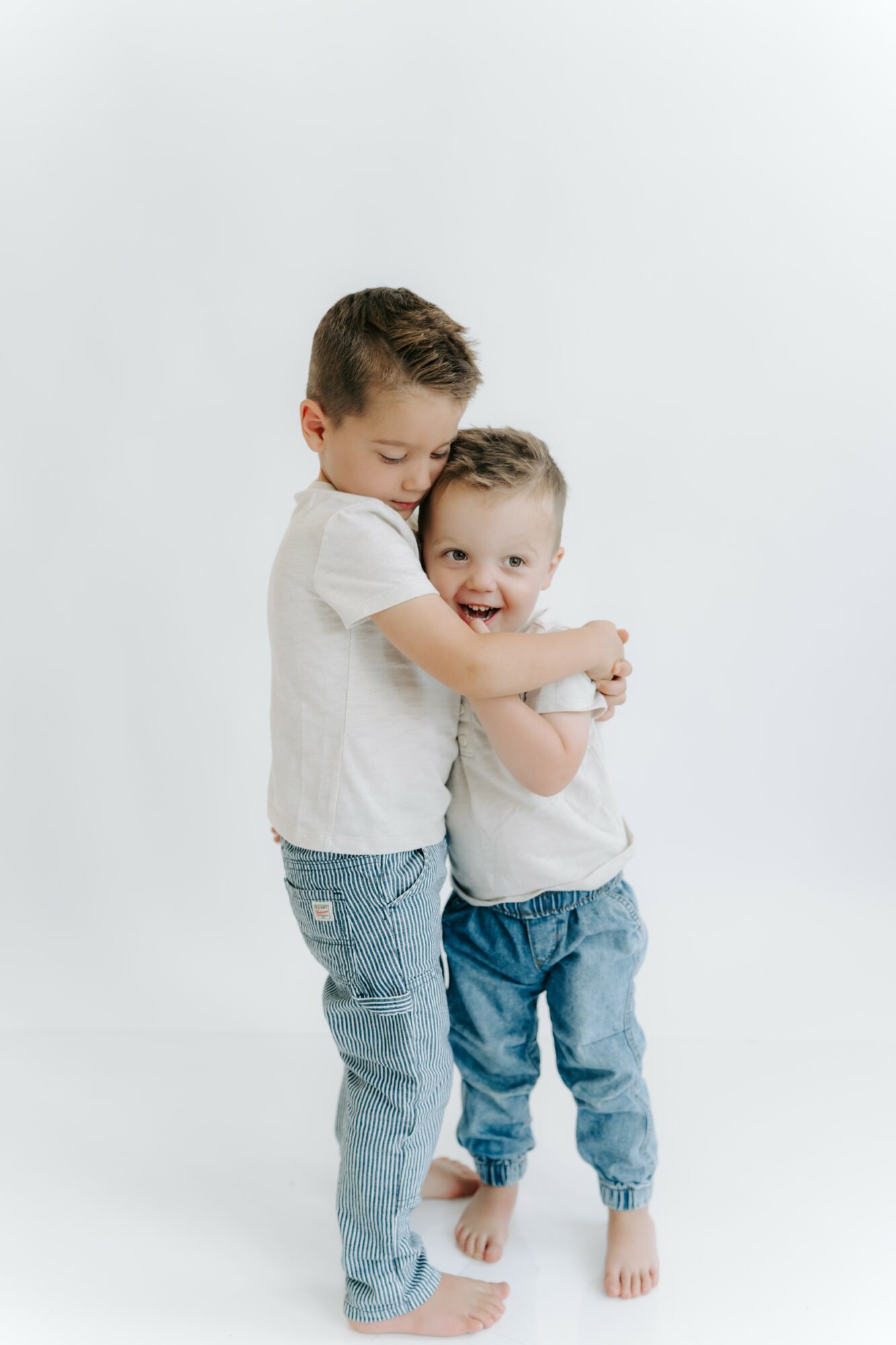 Two children hugging and smiling, one taller and older, the other younger, both wearing white shirts and jeans, standing barefoot.