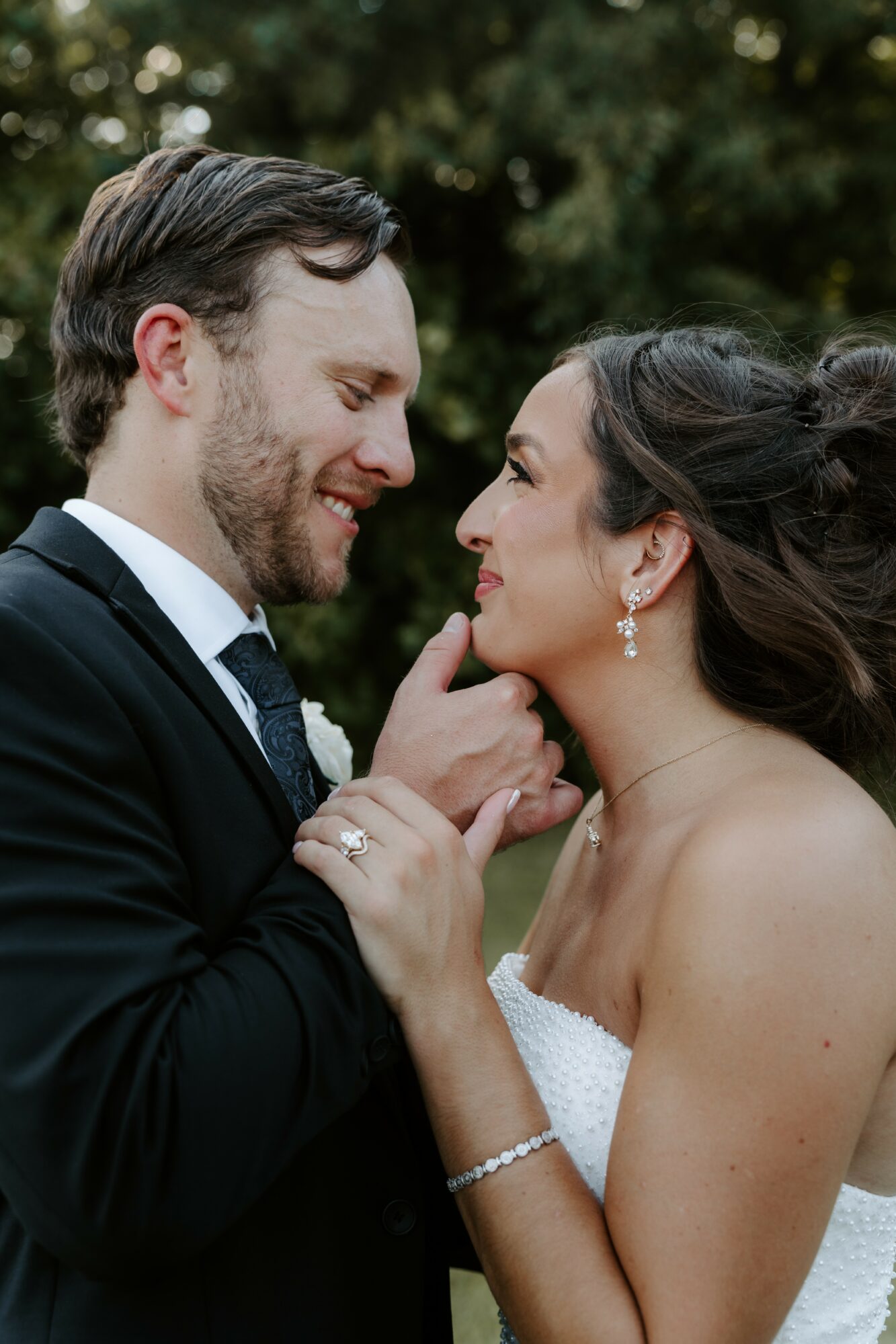 A couple in formal attire close together outdoors, gazing at each other affectionately, with trees in the background.