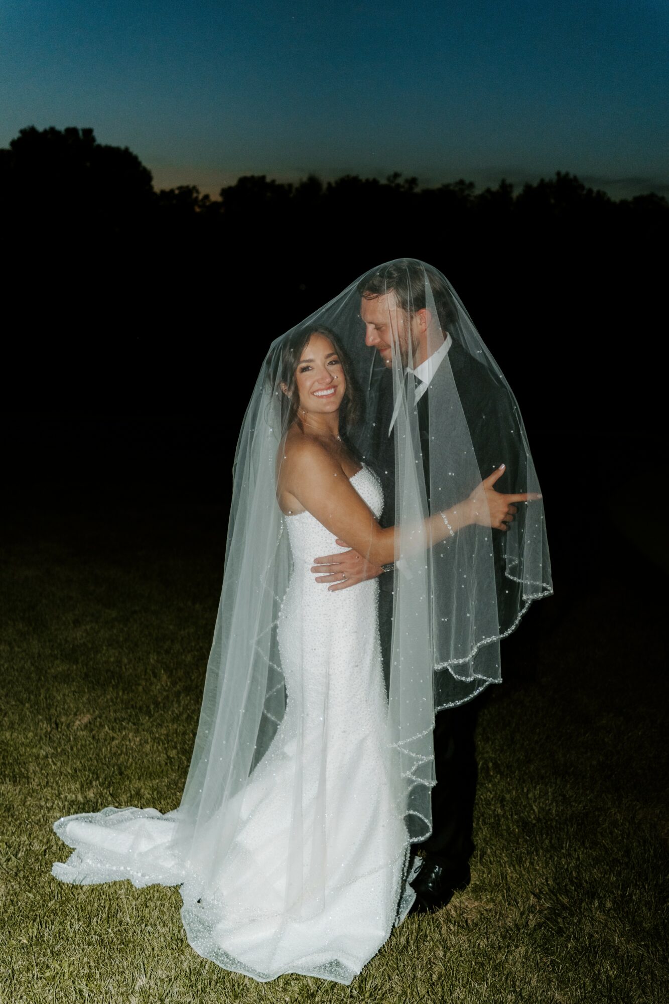 Bride and groom smiling and embracing outdoors at night, bride in white dress, groom in dark suit, veil covering both.
