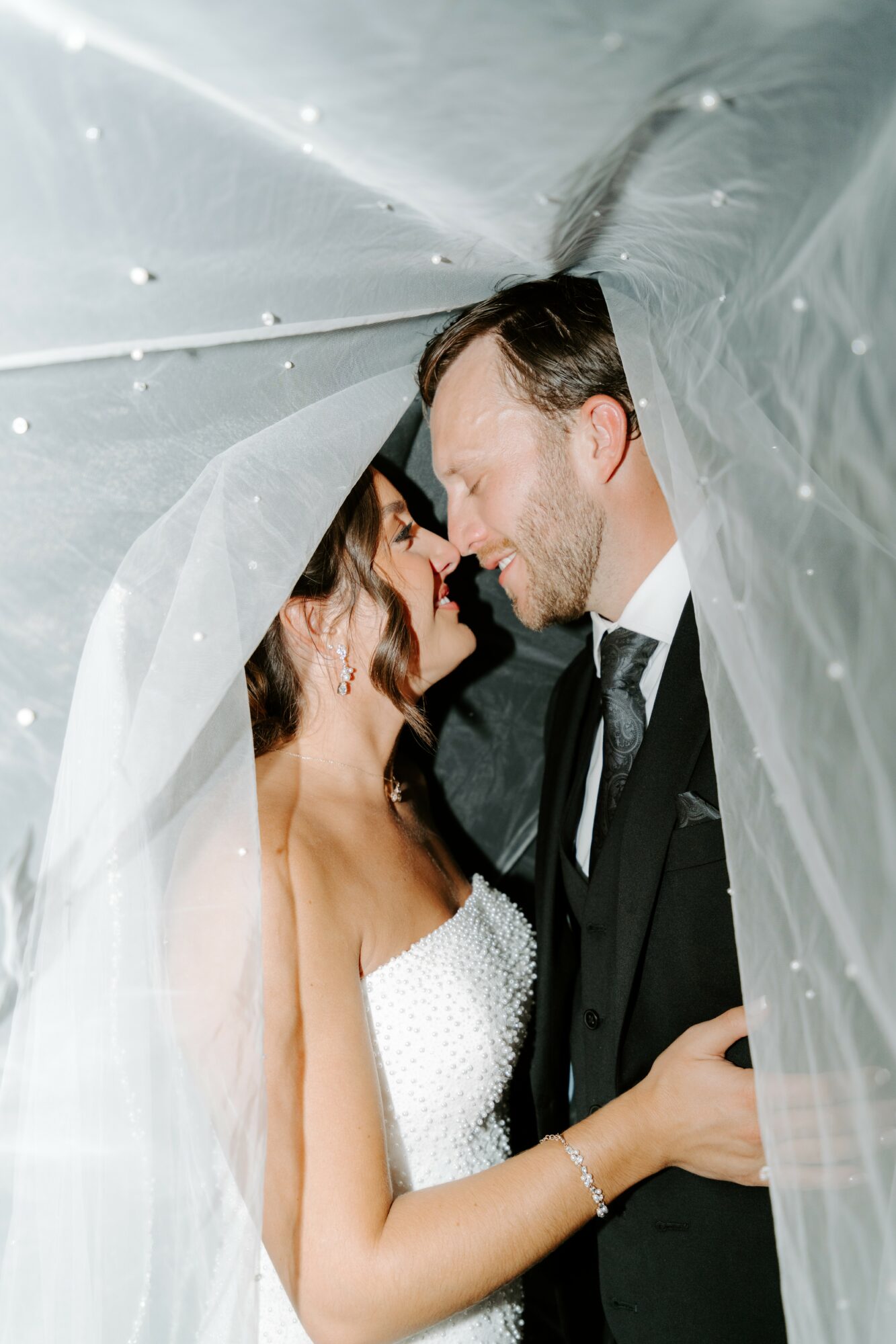 Bride and groom face each other under a veil, close and smiling, in wedding attire.