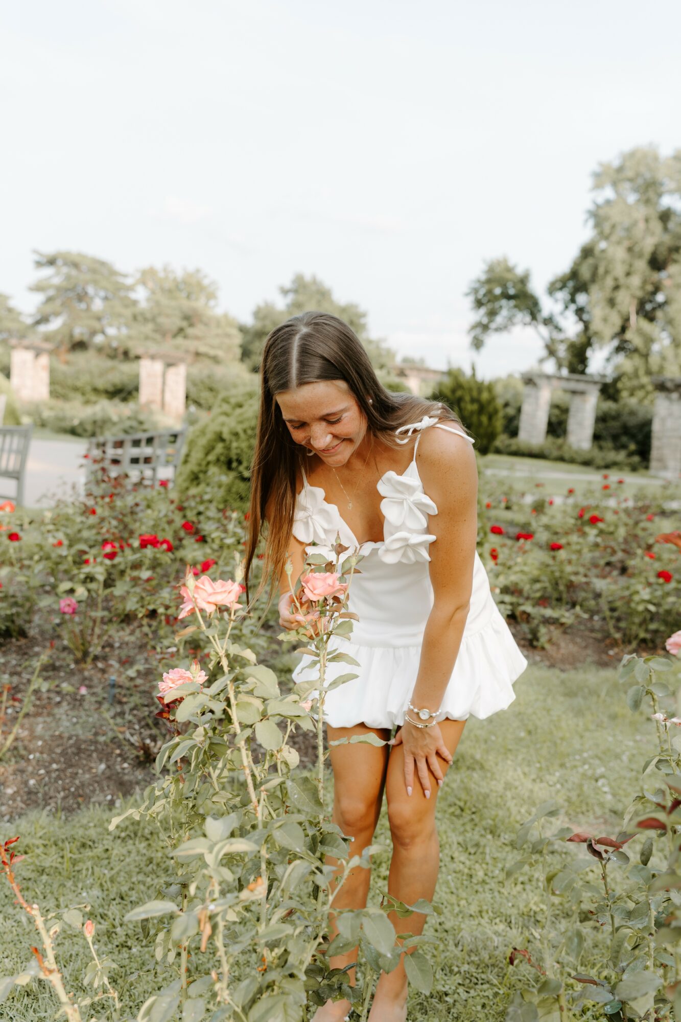 Young woman in white dress bending down in a garden with red flowers, trees, and a fence in the background.