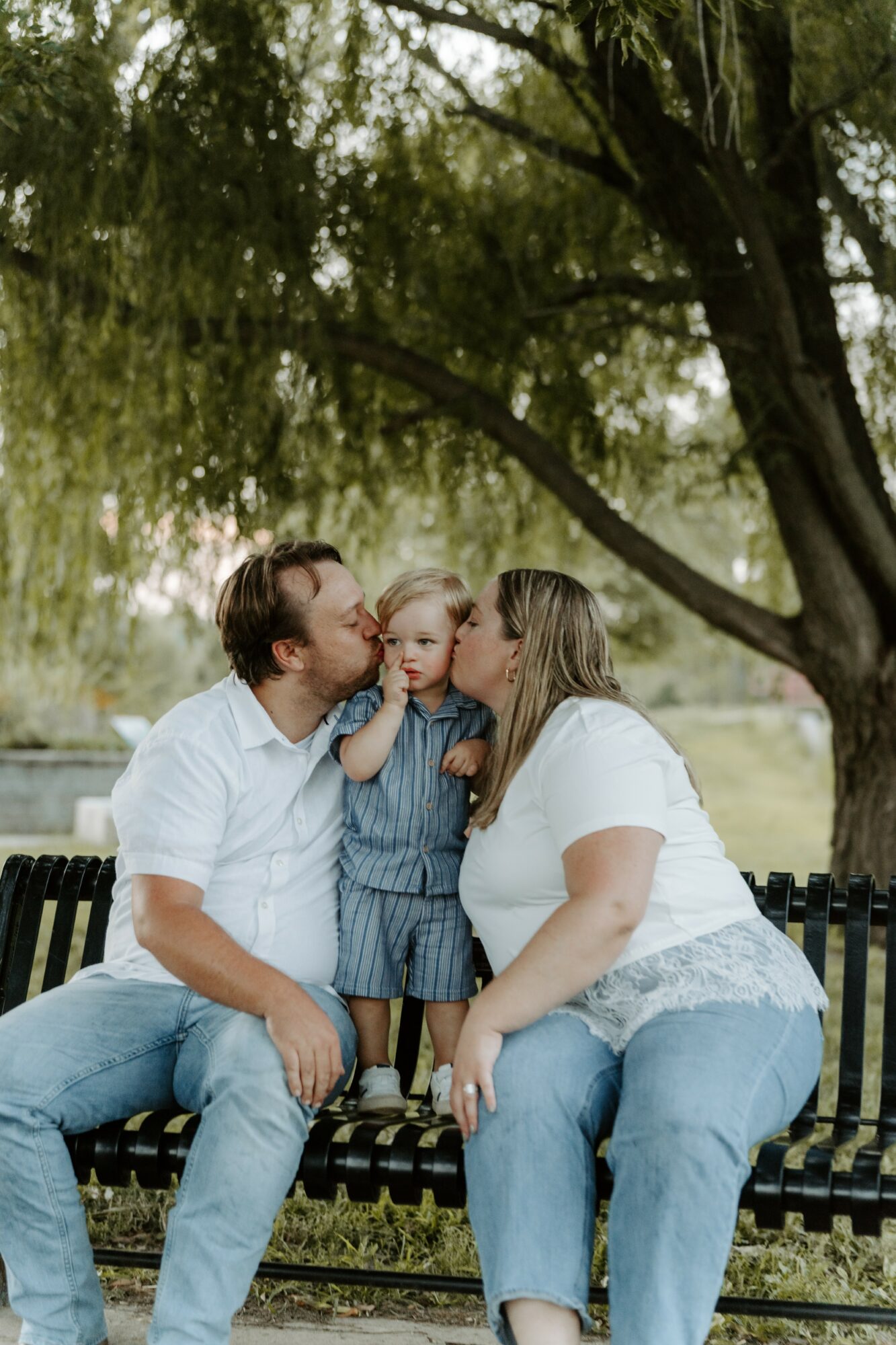 Family of three sitting on a park bench, with a large tree overhead, sharing a kiss and a smile.
