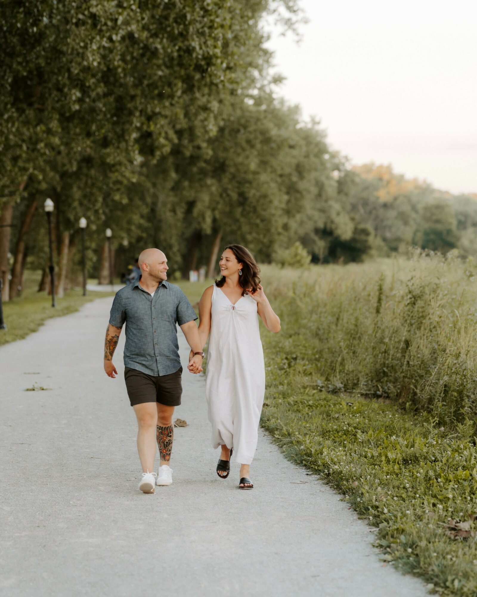 A couple walks hand in hand on a path surrounded by trees and grass, smiling at each other.