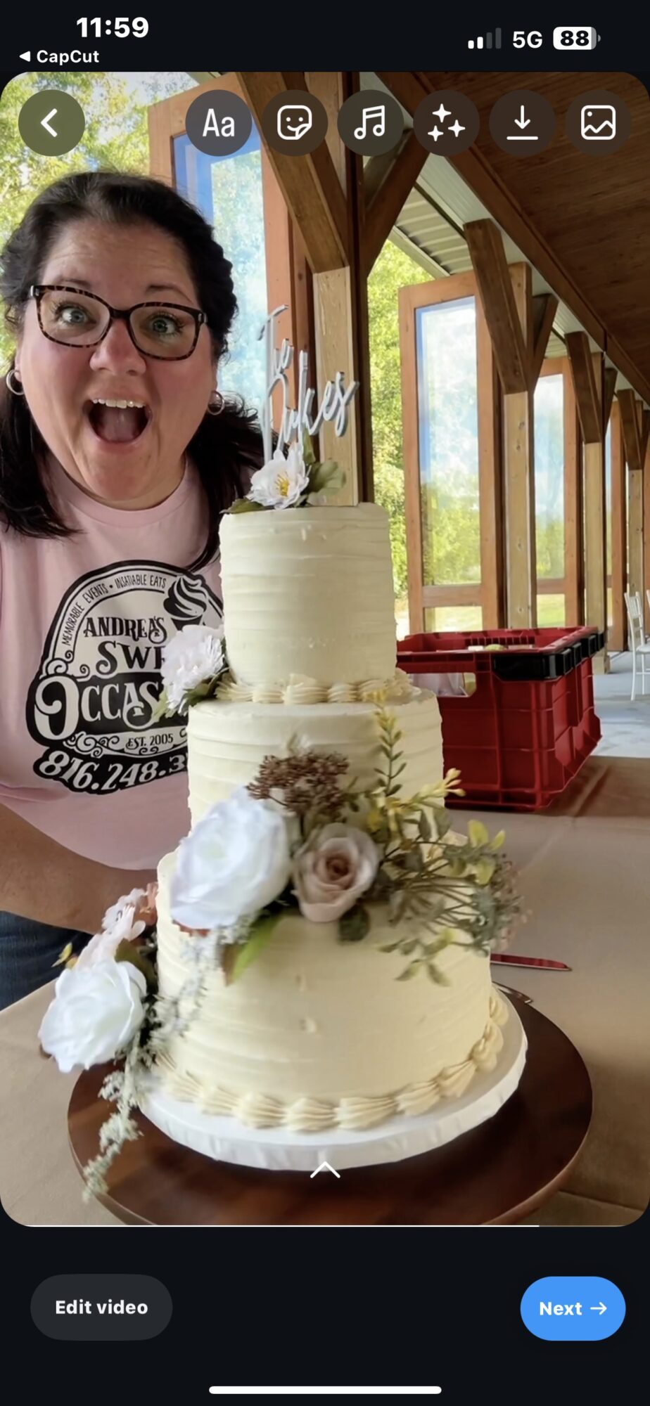 Woman with glasses smiling behind a two-tiered white wedding cake decorated with flowers, inside a wooden structure with large windows.