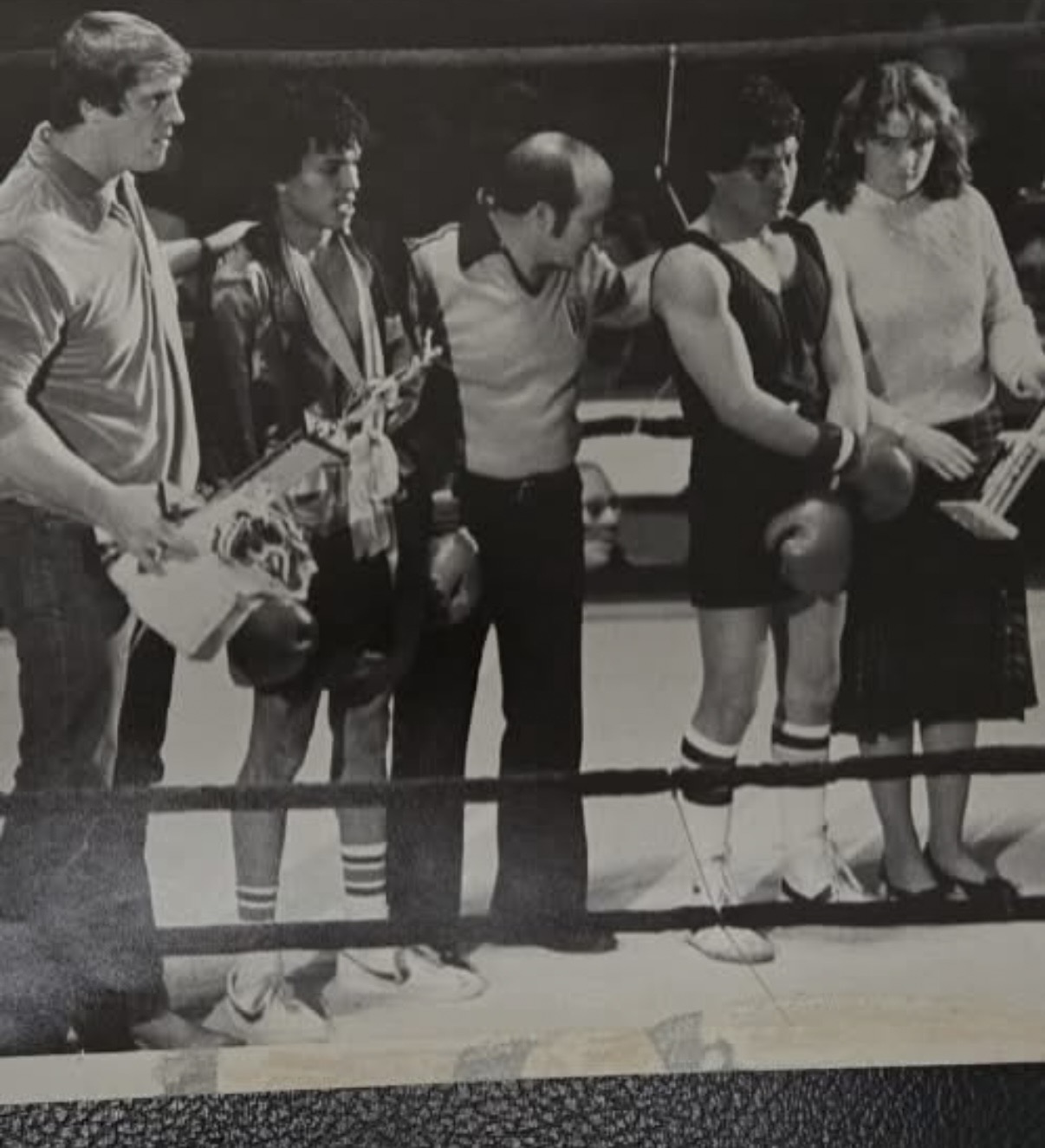 Six people stand in a boxing ring, some holding boxing gloves and equipment, in black and white. Word count: 20.