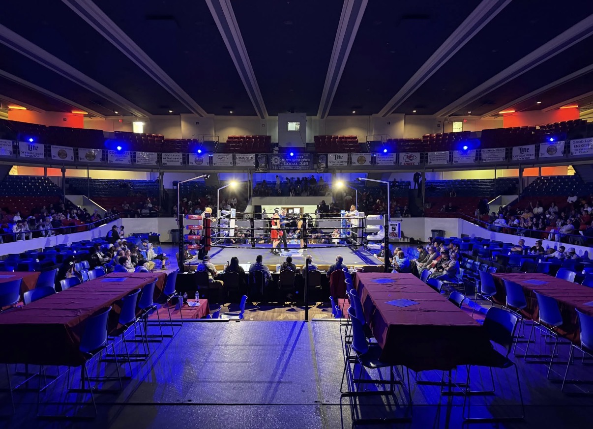 Boxing ring in center of arena with seating and tables around, illuminated by colorful lights, audience seated on multiple levels.
