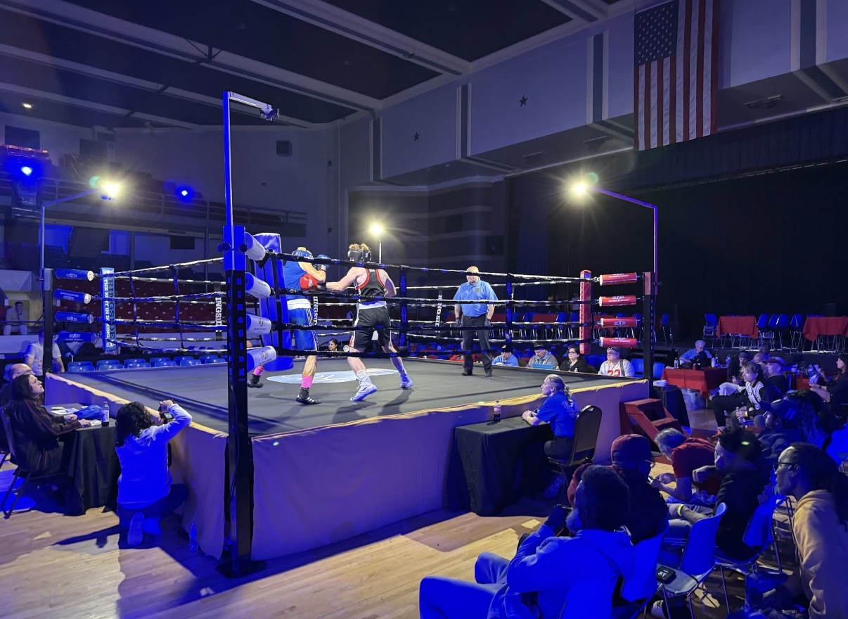 Boxers in a ring sparring, referee watching, audience seated around the ring, indoor event with bright lighting.