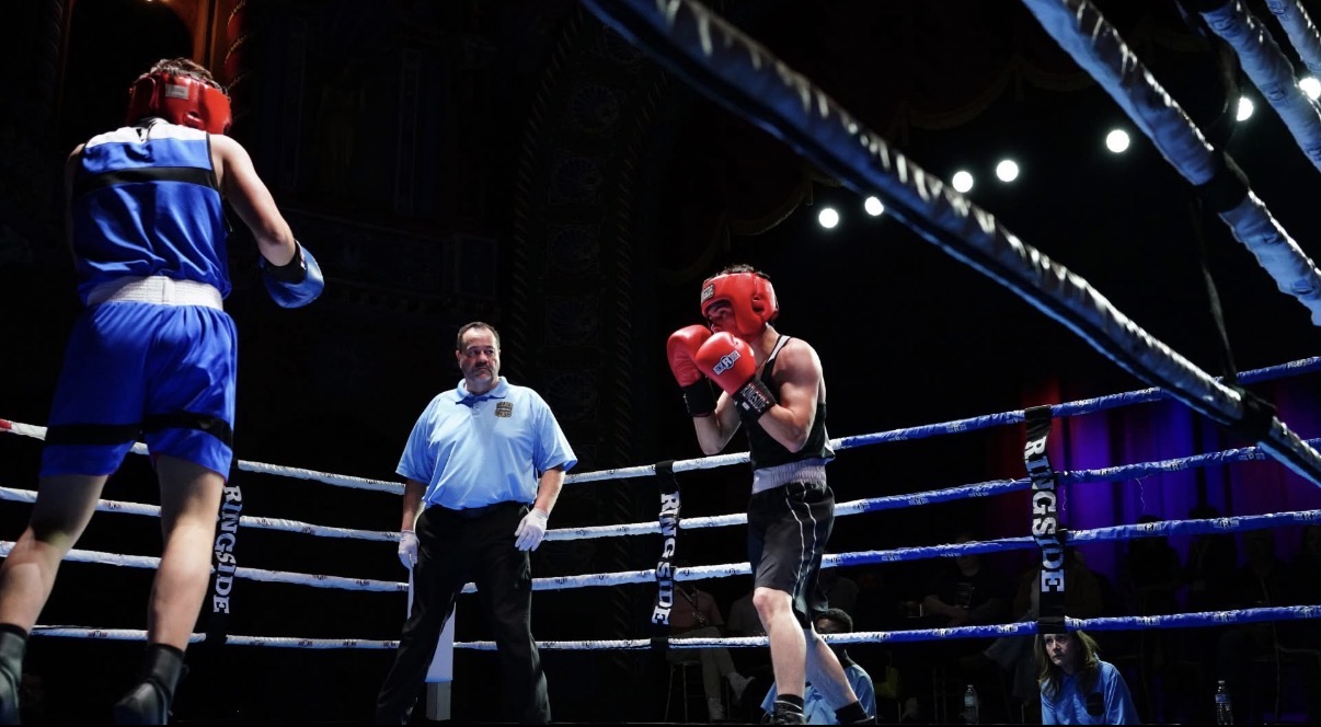 Two boxers in a ring with a referee, one boxer in red gloves and the other in blue, under bright lights.
