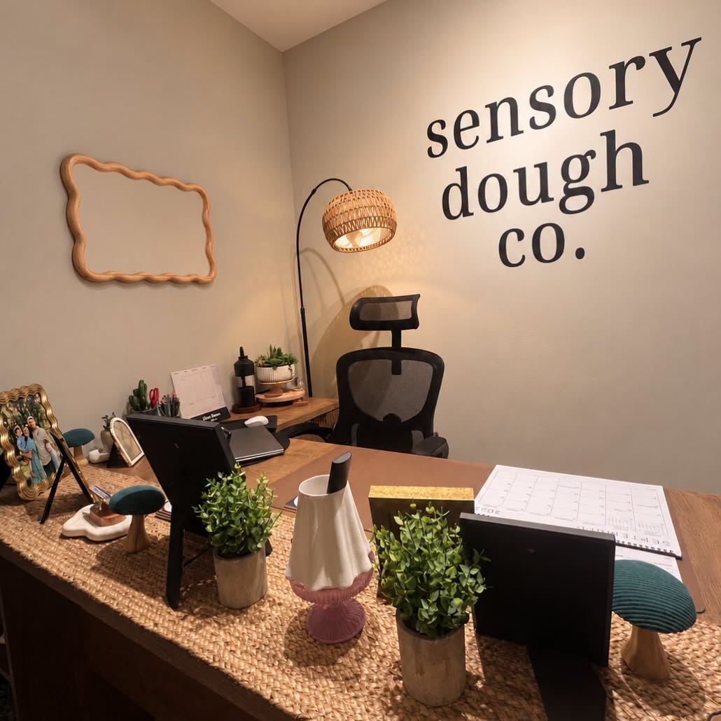 Office desk with plants, a lamp, and office supplies, against a wall with the text 'sensory dough co.'