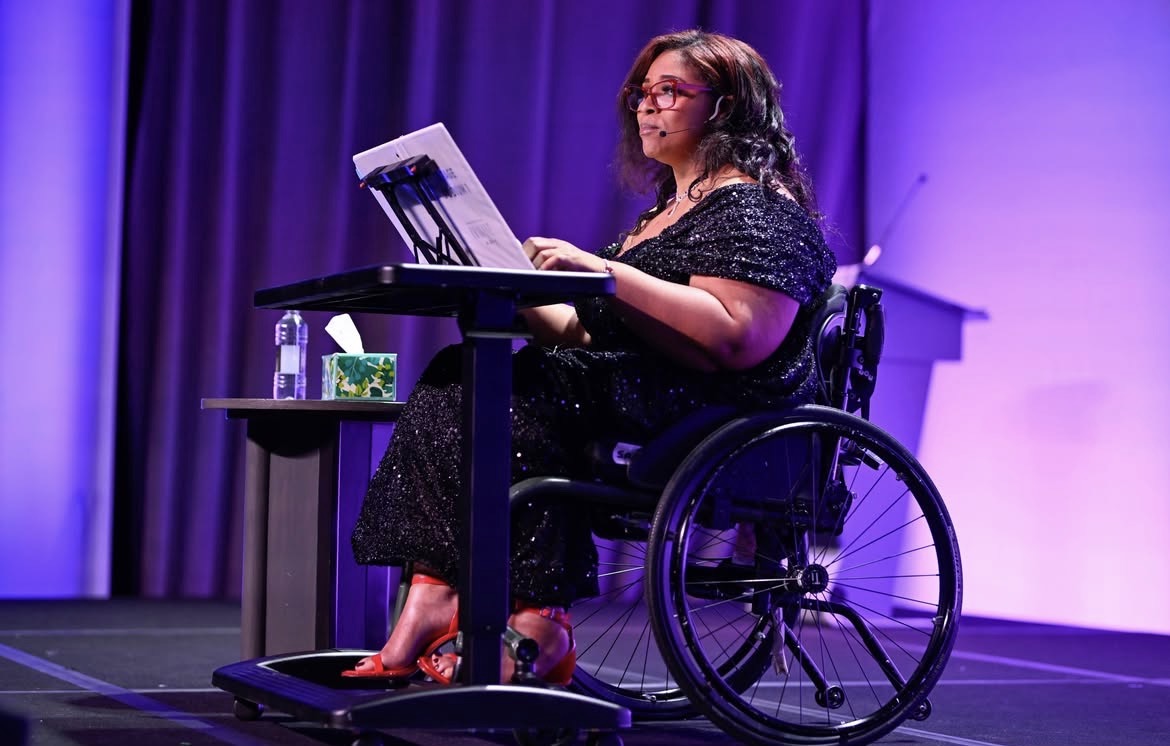 Woman in wheelchair playing a keyboard on stage with purple lighting, wearing glasses and a black dress.