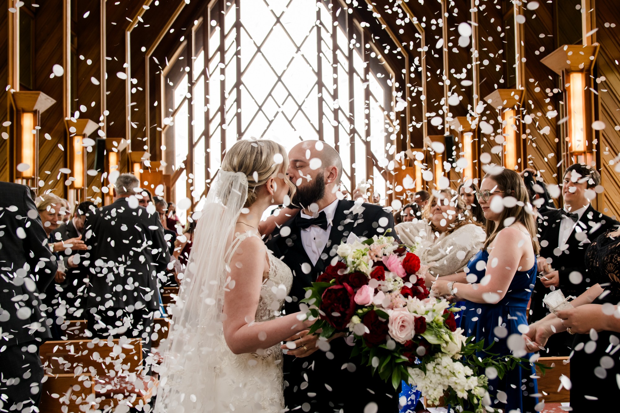 Bride and groom kiss under confetti in a decorated wedding venue with guests celebrating, holding flowers and confetti, bright lighting.