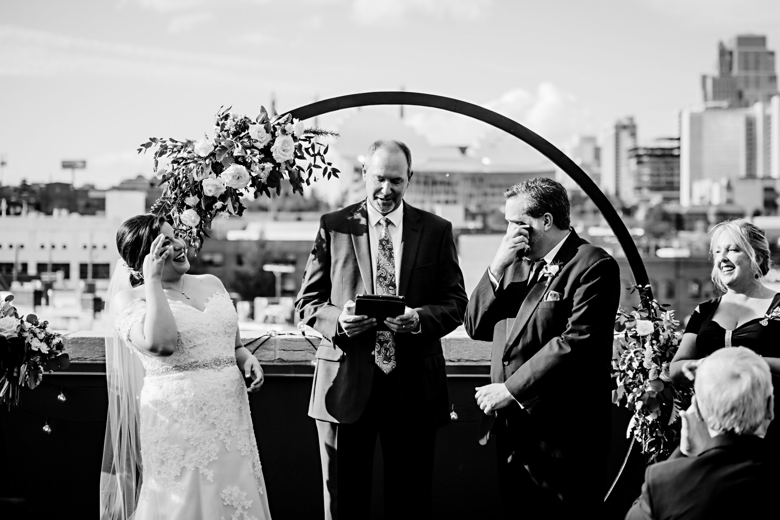 Group of people at a wedding ceremony outdoors, with city buildings in the background, including a bride, groom, and officiant.