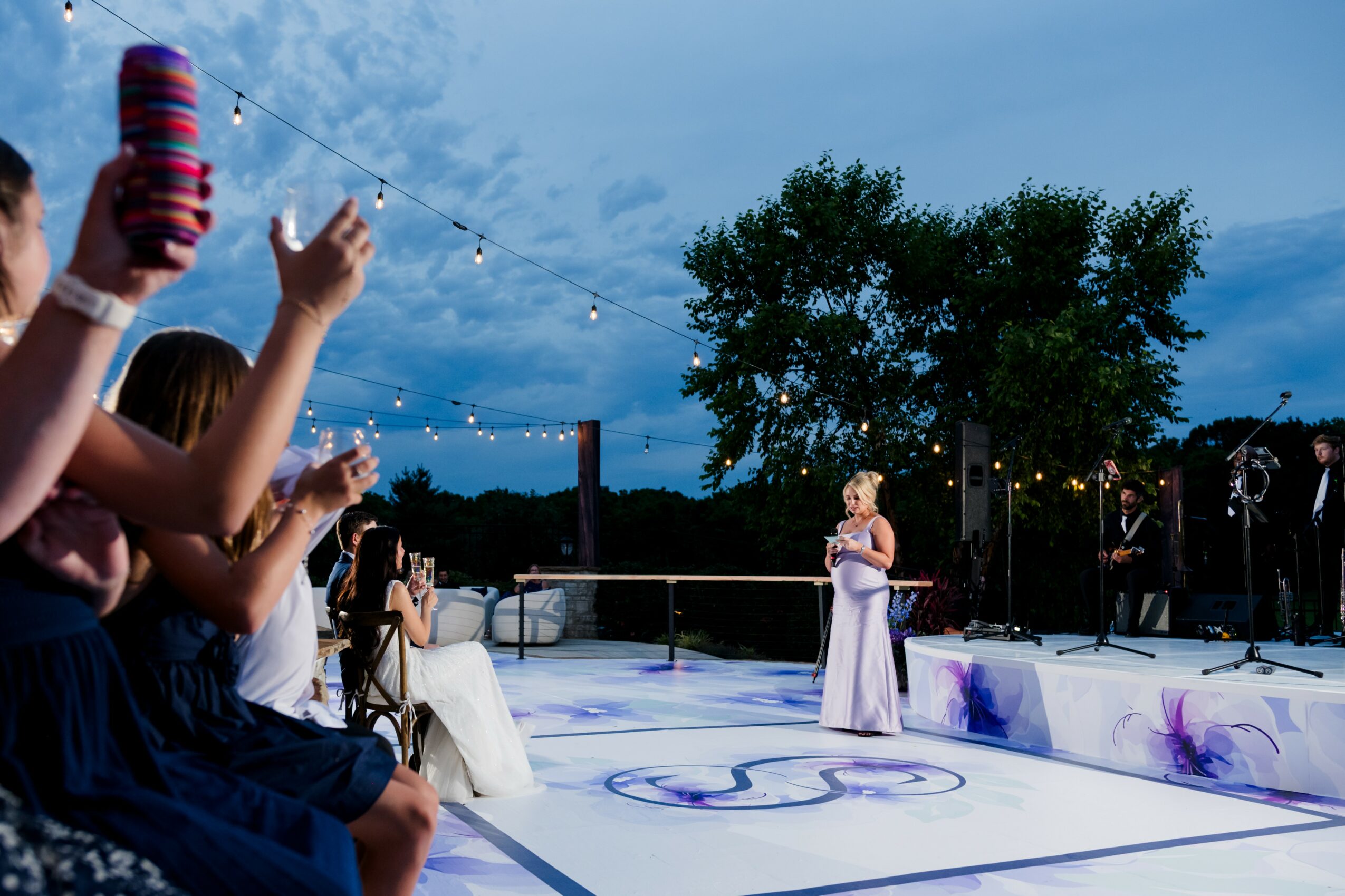 A woman in a white dress stands on an outdoor stage during evening, with trees and string lights in the background.