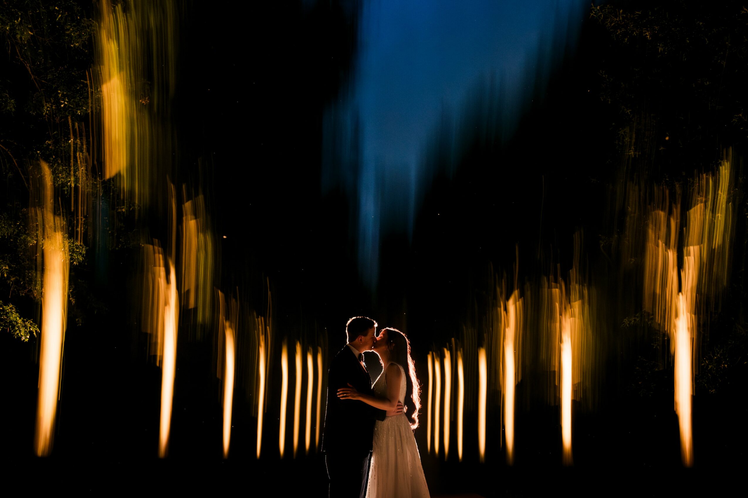 A couple embraces in a dark outdoor setting with illuminated trees and a blue sky above, backlit to create a silhouette effect.