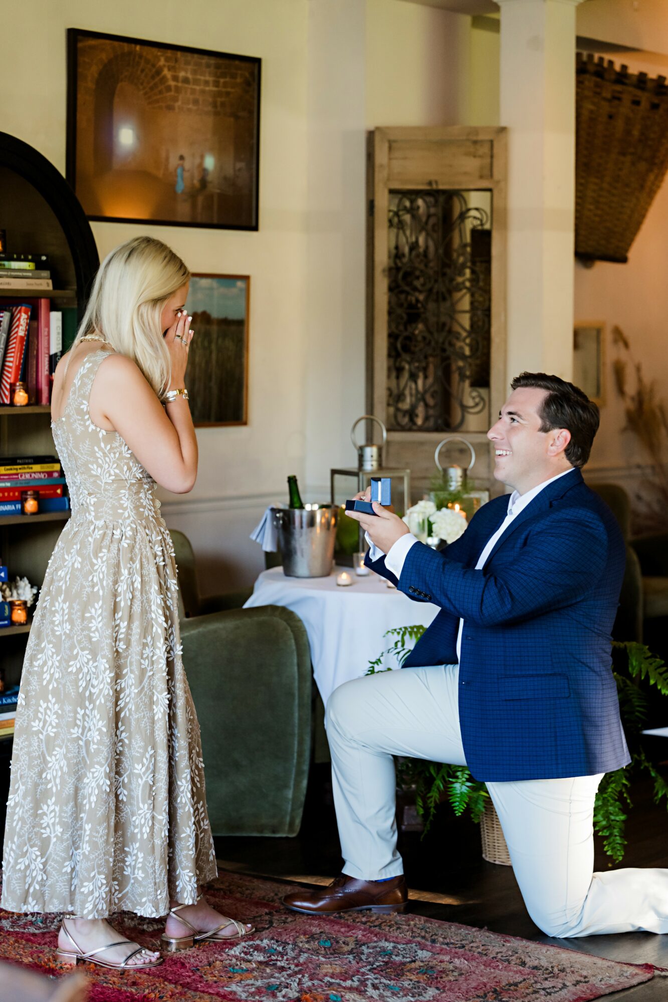 Man kneeling with a ring box, woman in a dress reacting happily, indoor setting with artwork and furniture.