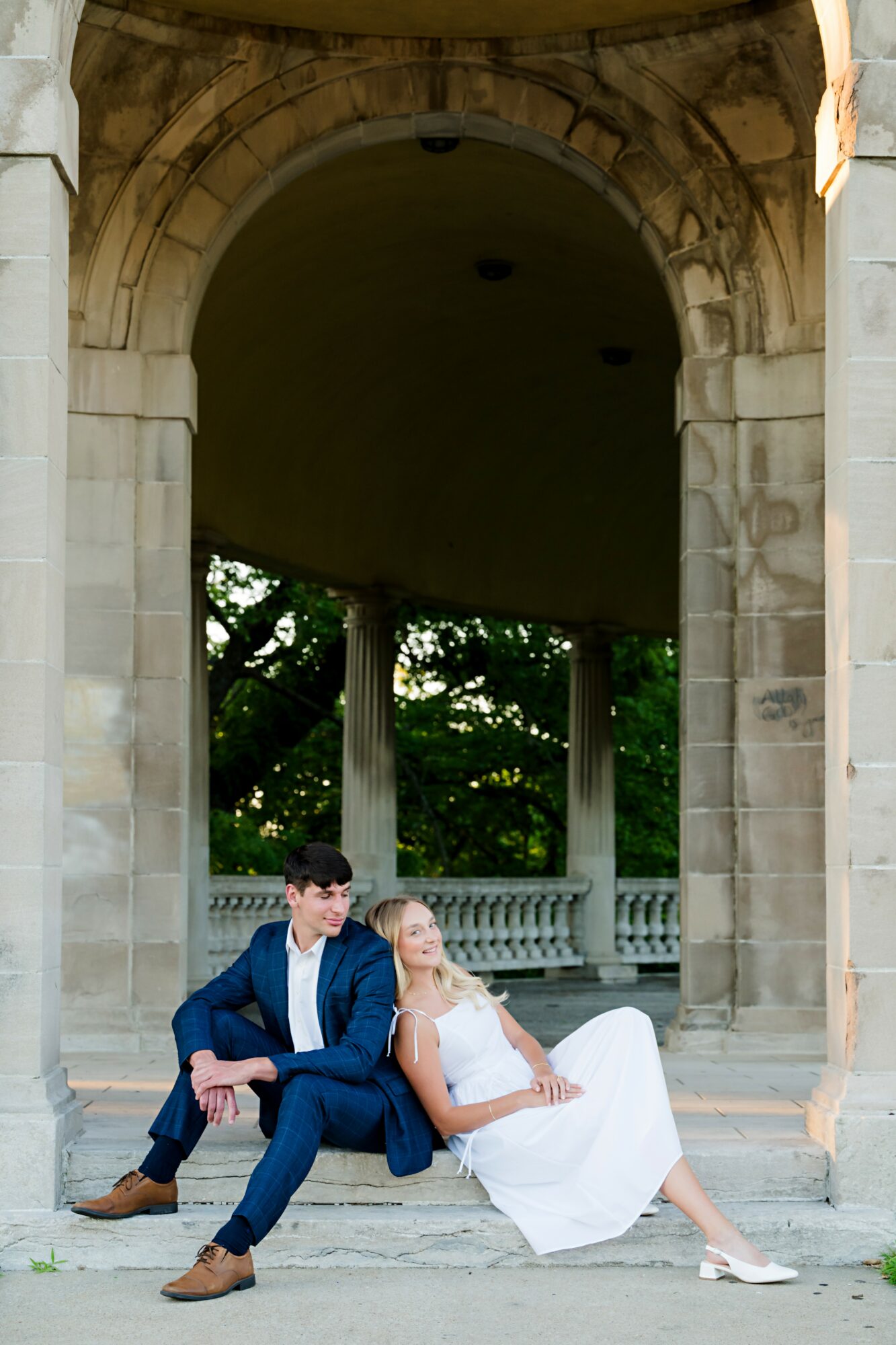 A couple in wedding attire sitting on steps under an archway with trees in the background.