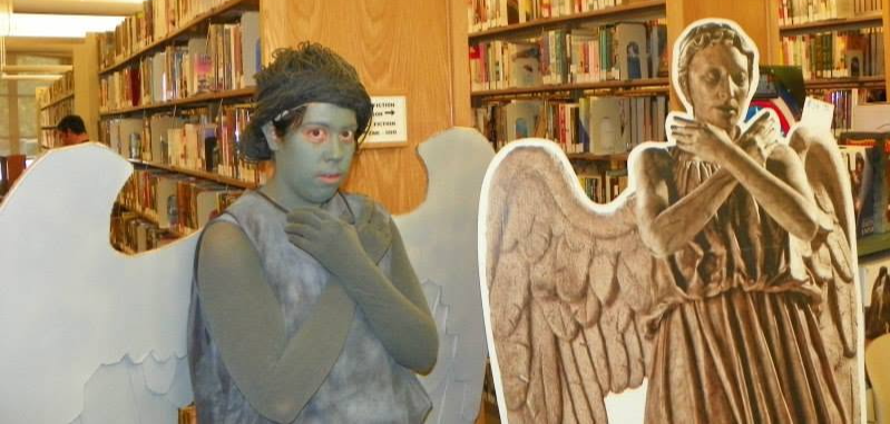 Two people dressed as angels with wings in a library, one with a headband and the other with a feathered costume.