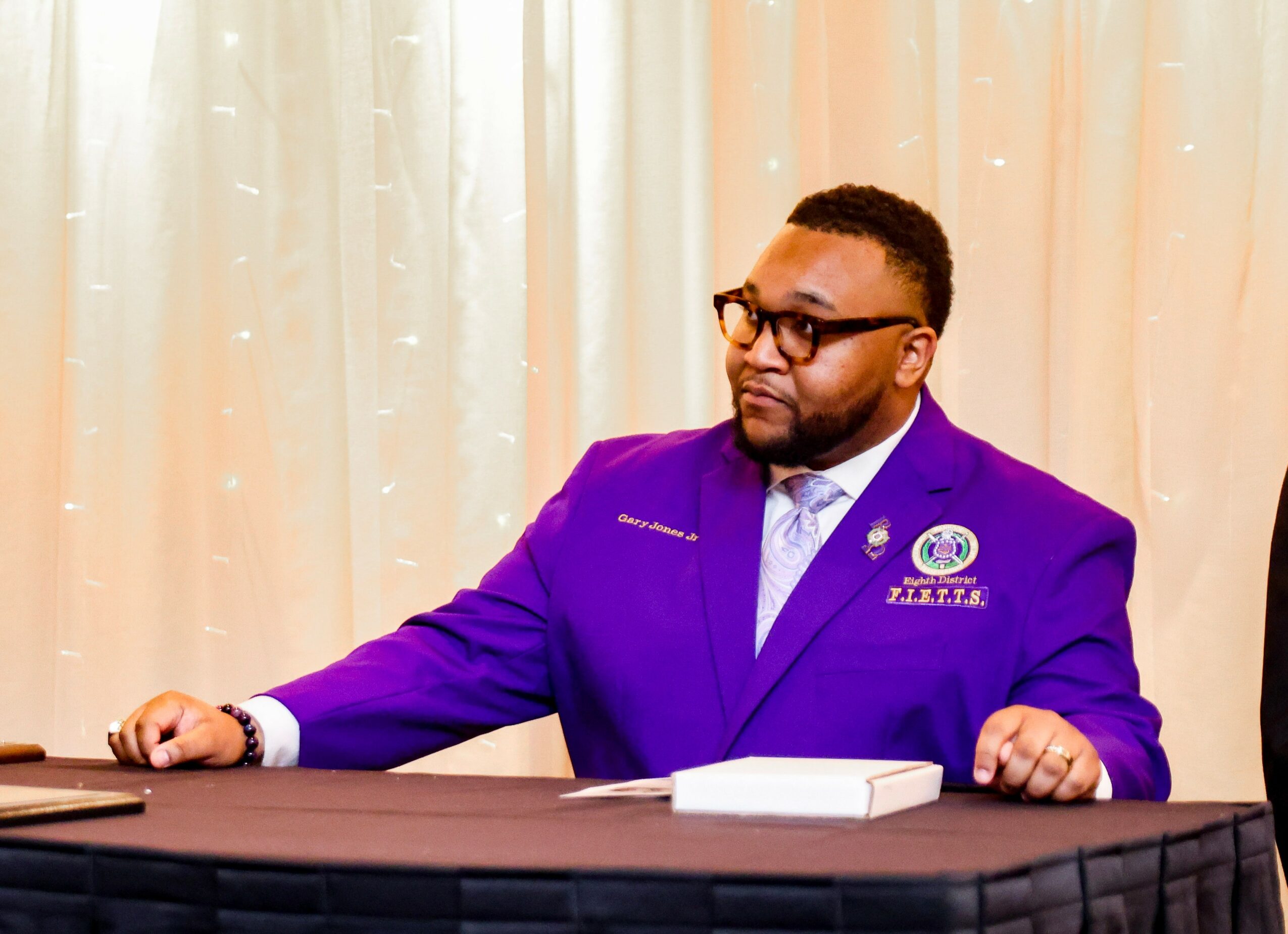 Man in purple suit sitting at a table with a curtain background, looking to the side, with a notebook in front.