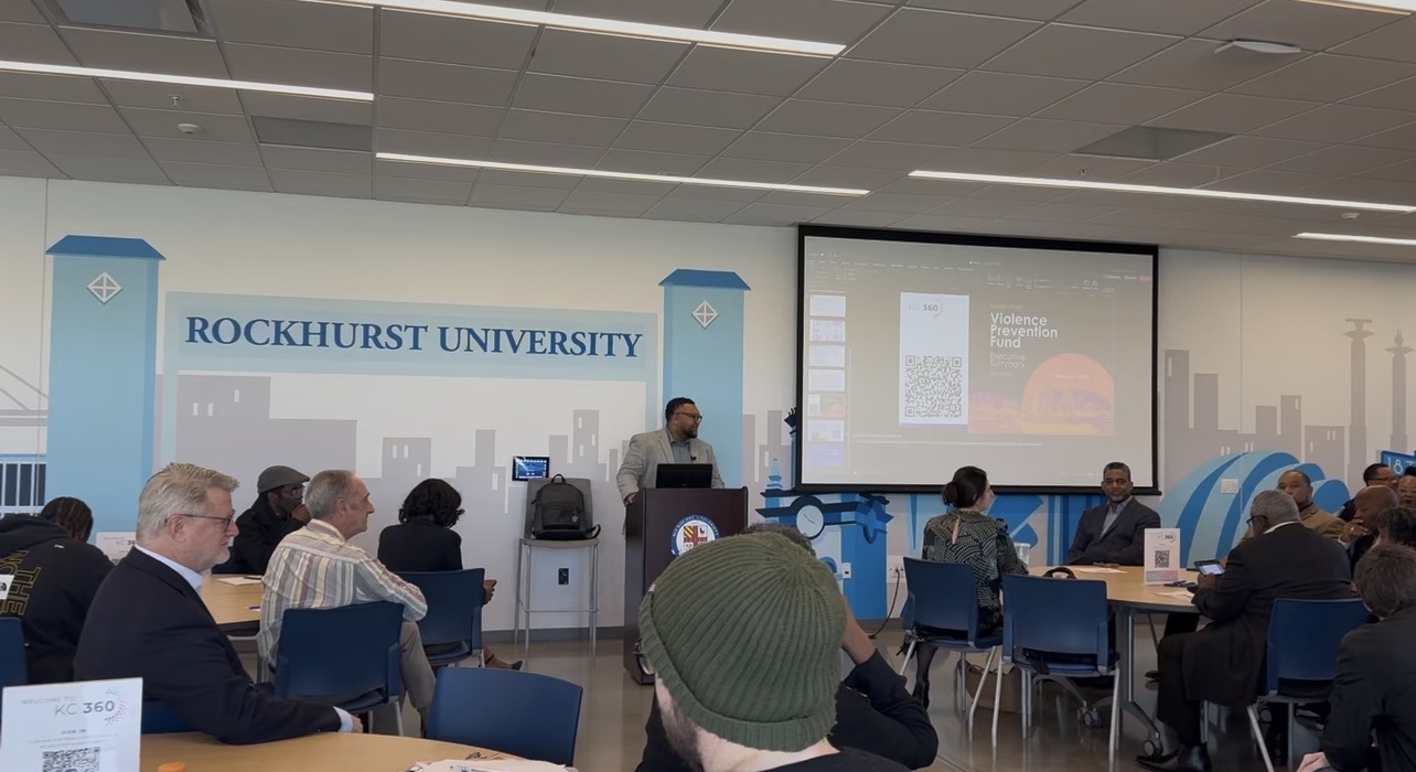 Lecture hall with a speaker at podium, audience seated, presentation screen displaying slides, and a banner reading 'ROCKHURST UNIVERSITY'.