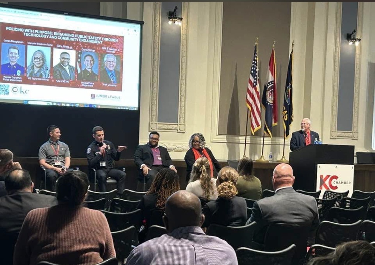 Panel discussion with five people on stage, audience seated, flags and a large screen displaying images behind them.