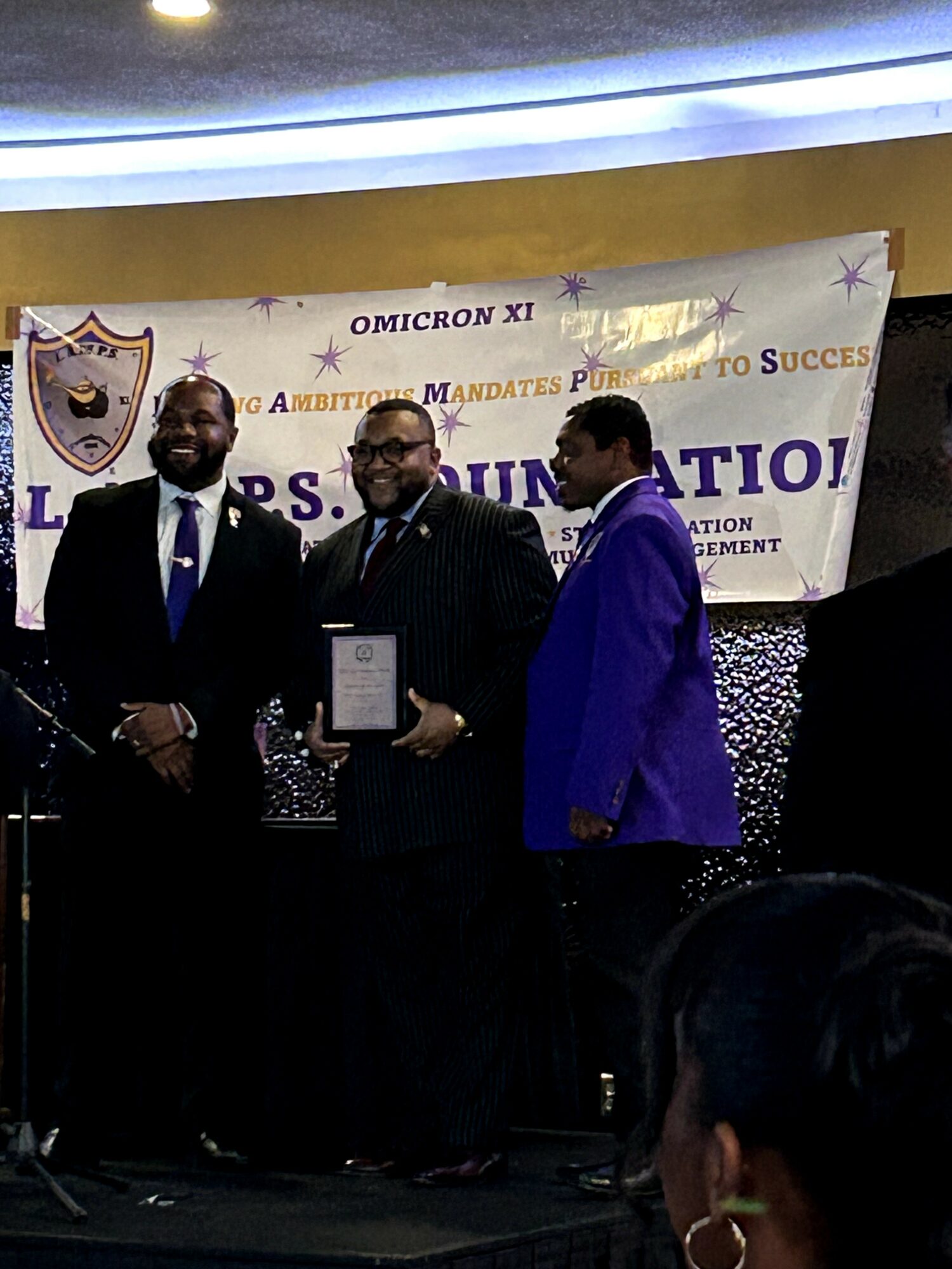 Three men in formal suits standing on stage at an event, with a banner behind them reading 'Omonicon XI' and 'Celebration'.