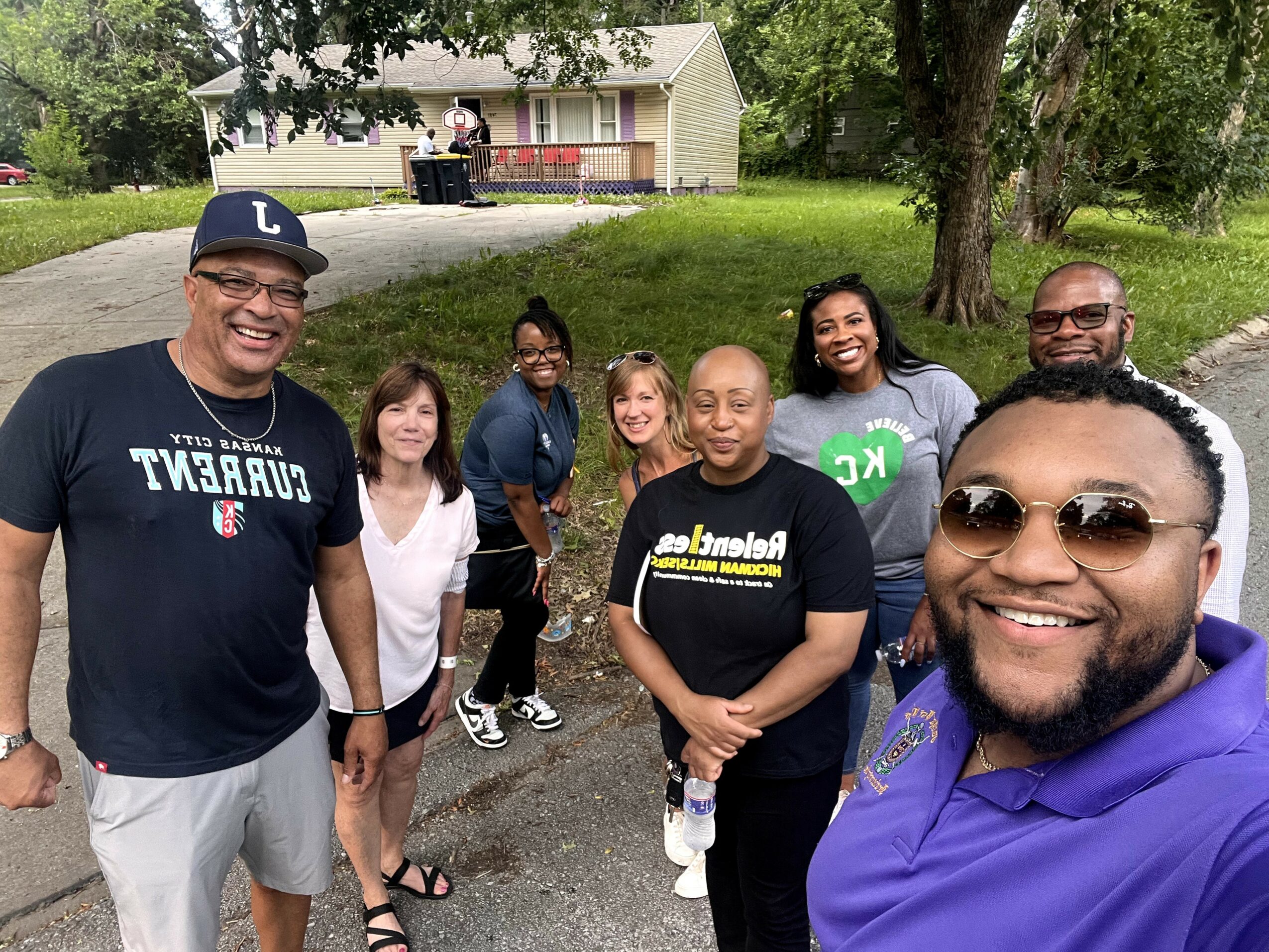 Group of eight people outdoors on a sidewalk, smiling, with trees and a house in the background.