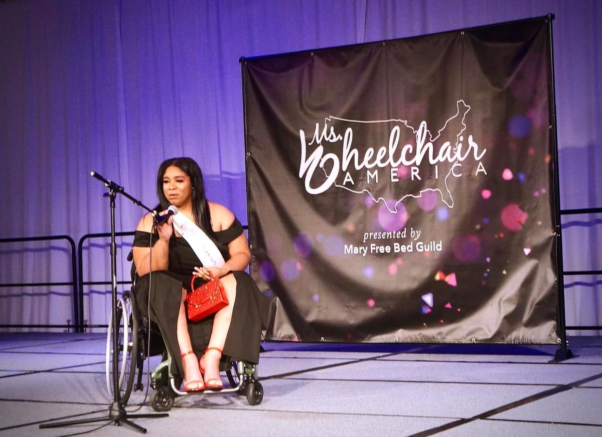 Woman in wheelchair on stage with microphone, backdrop reads 'Miss Wheelchair America,' purple lighting, holding a red bag.